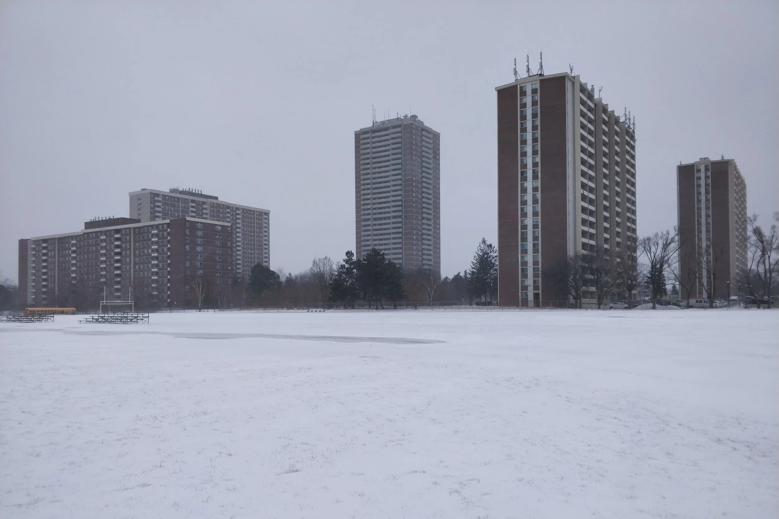 A snow-covered field with three tall apartment buildings in the background, under a gray overcast sky.