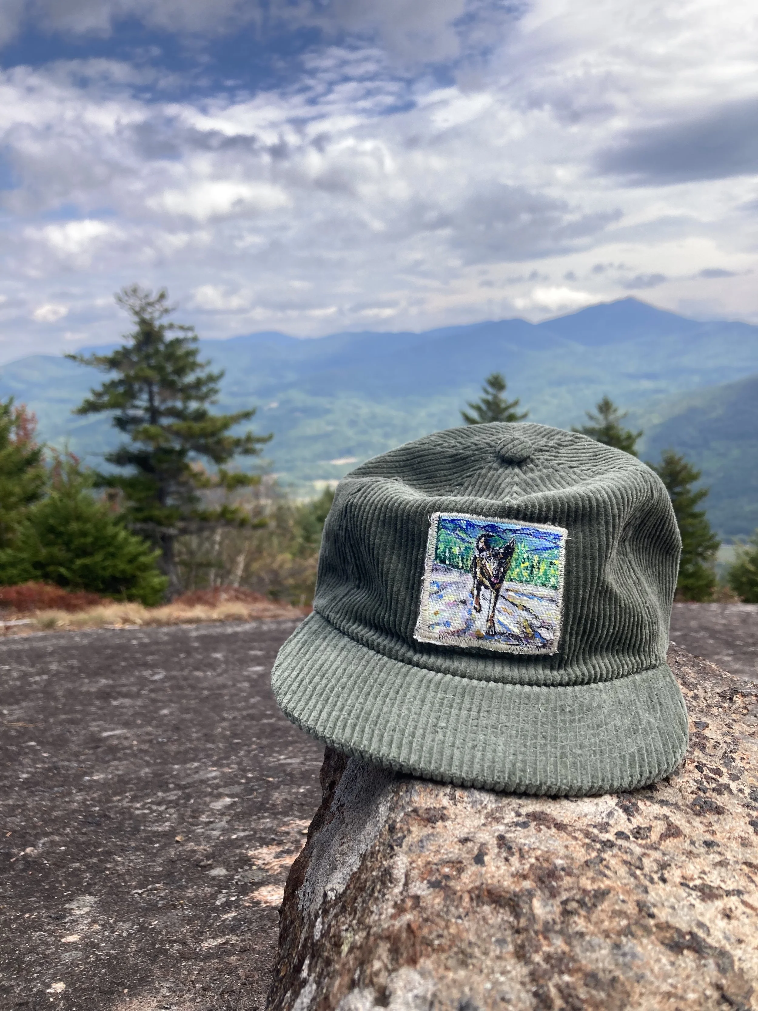Green corduroy bucket hat with embroidered patch of a dog, set on a rock with a mountain and cloudy sky in the background.