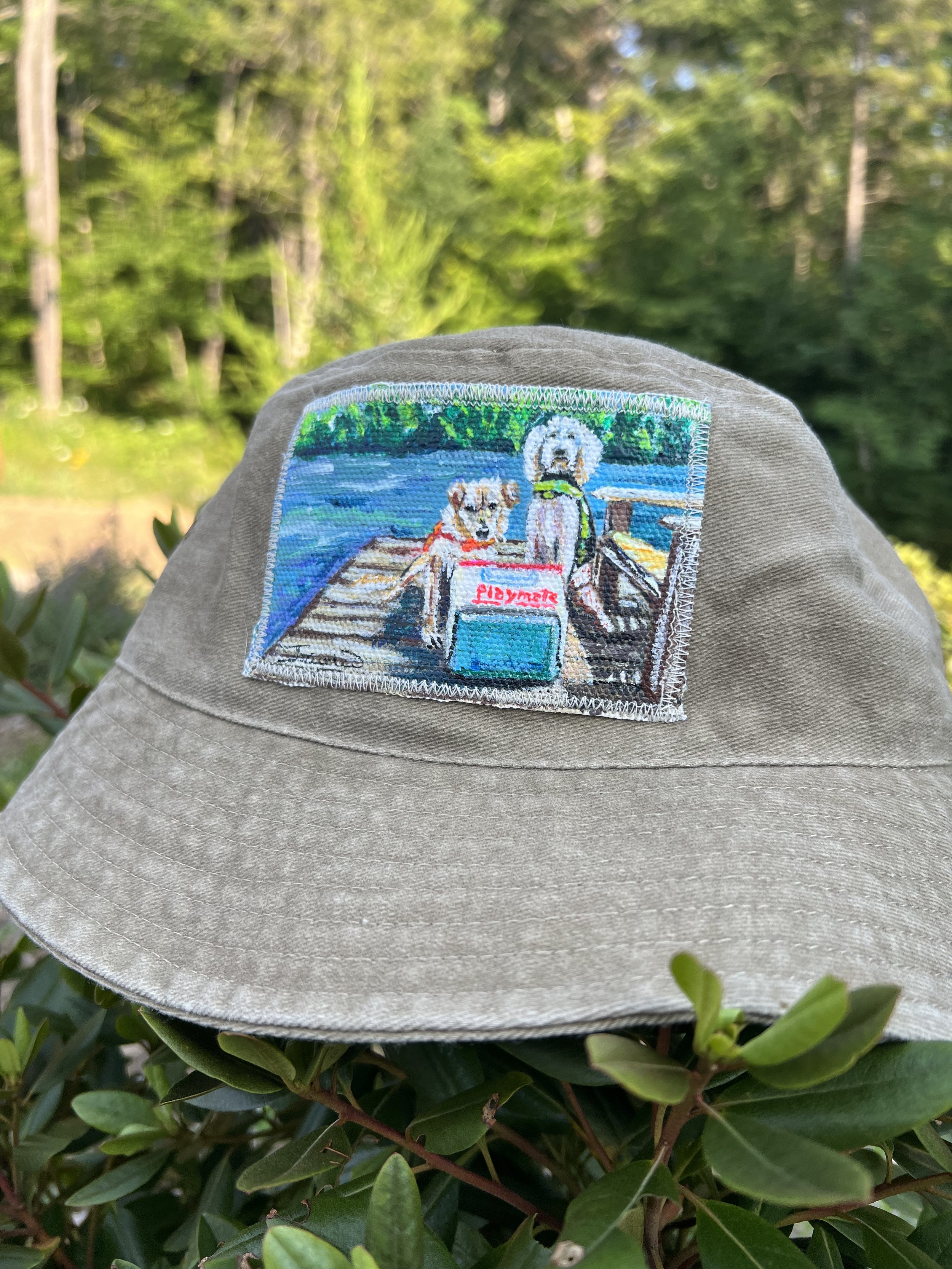 A beige bucket hat with an embroidered patch of two dogs sitting on a dock by a lake, with trees in the background.