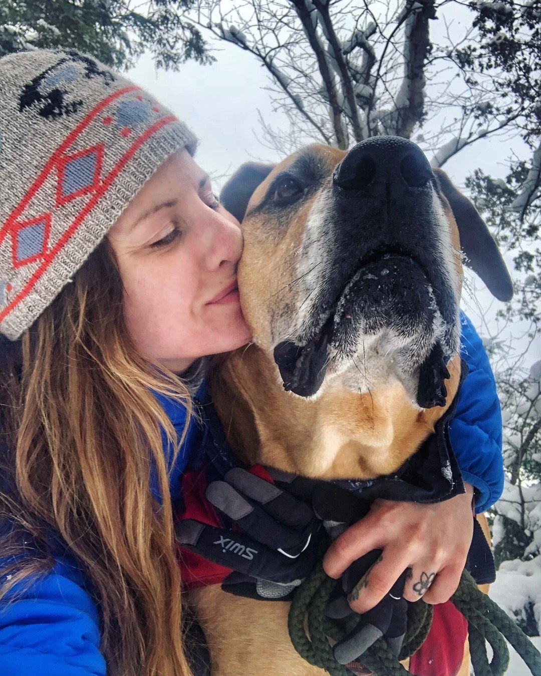 A young woman with long red hair, wearing a gray beanie with colorful patterns, hugging and kissing a large brown and black dog in a snowy outdoor setting with snow-covered trees in the background.