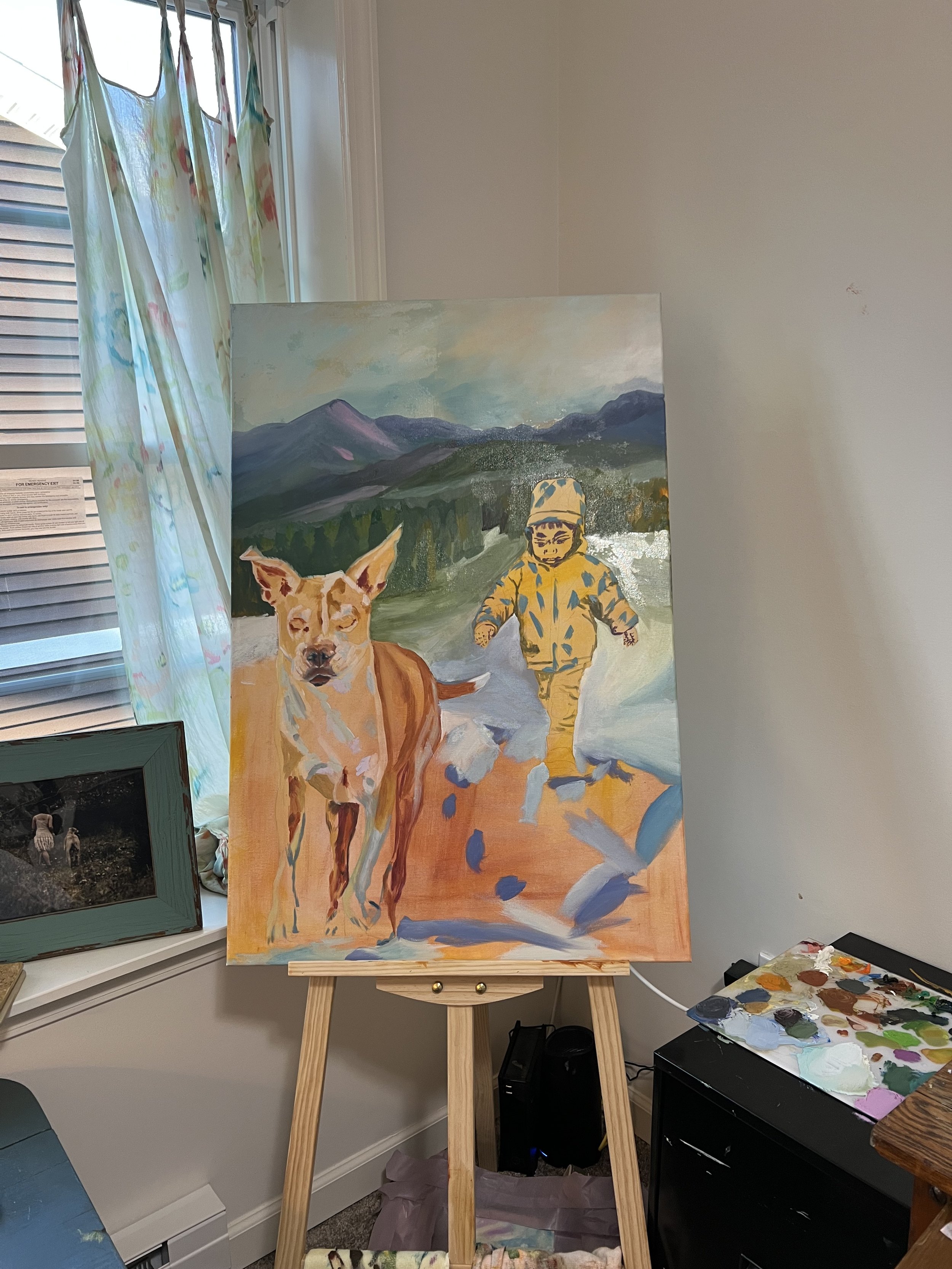 An unfinished painting on an easel depicts a child in an orange and yellow snowsuit and a dog walking in a mountainous landscape with a cloudy sky. The room has a window with floral curtains, a framed photo on the windowsill, and a palette with mixed