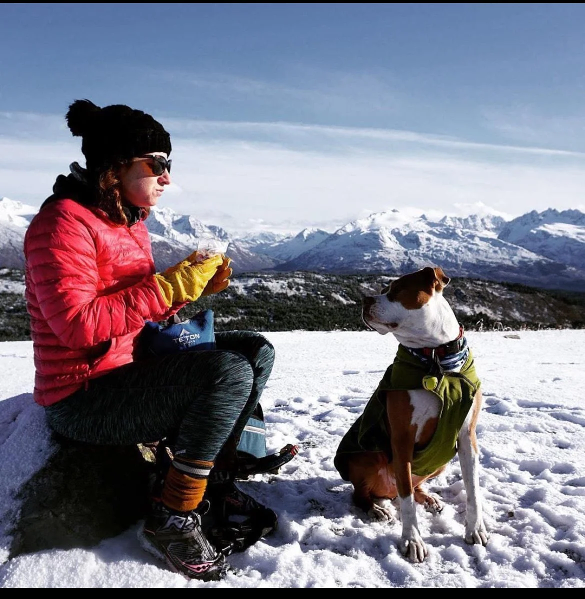 A woman in winter gear, holding a snack, sitting on snow with mountains in the background; a dog wearing a green jacket sitting nearby in a snowy landscape.