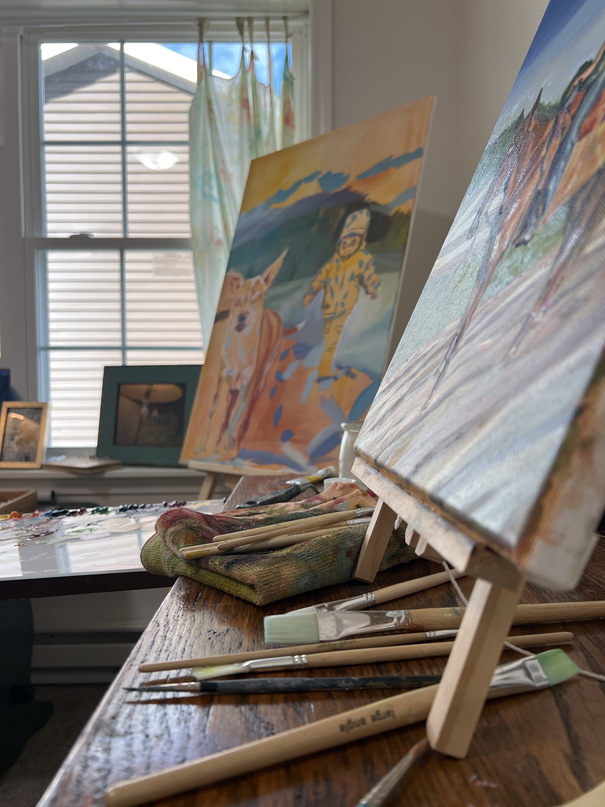 View of an art studio with two small landscape paintings on easels, paintbrushes, and a palette on a wooden table, with a window in the background showing sunny weather and a house's siding.