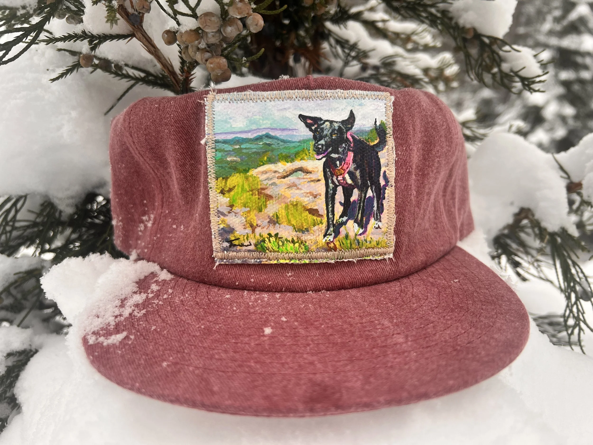 A red baseball cap with a patch of a black dog standing on a landscape with green hills, blue sky, and colorful vegetation, placed on snow and surrounded by snow-covered pine branches.