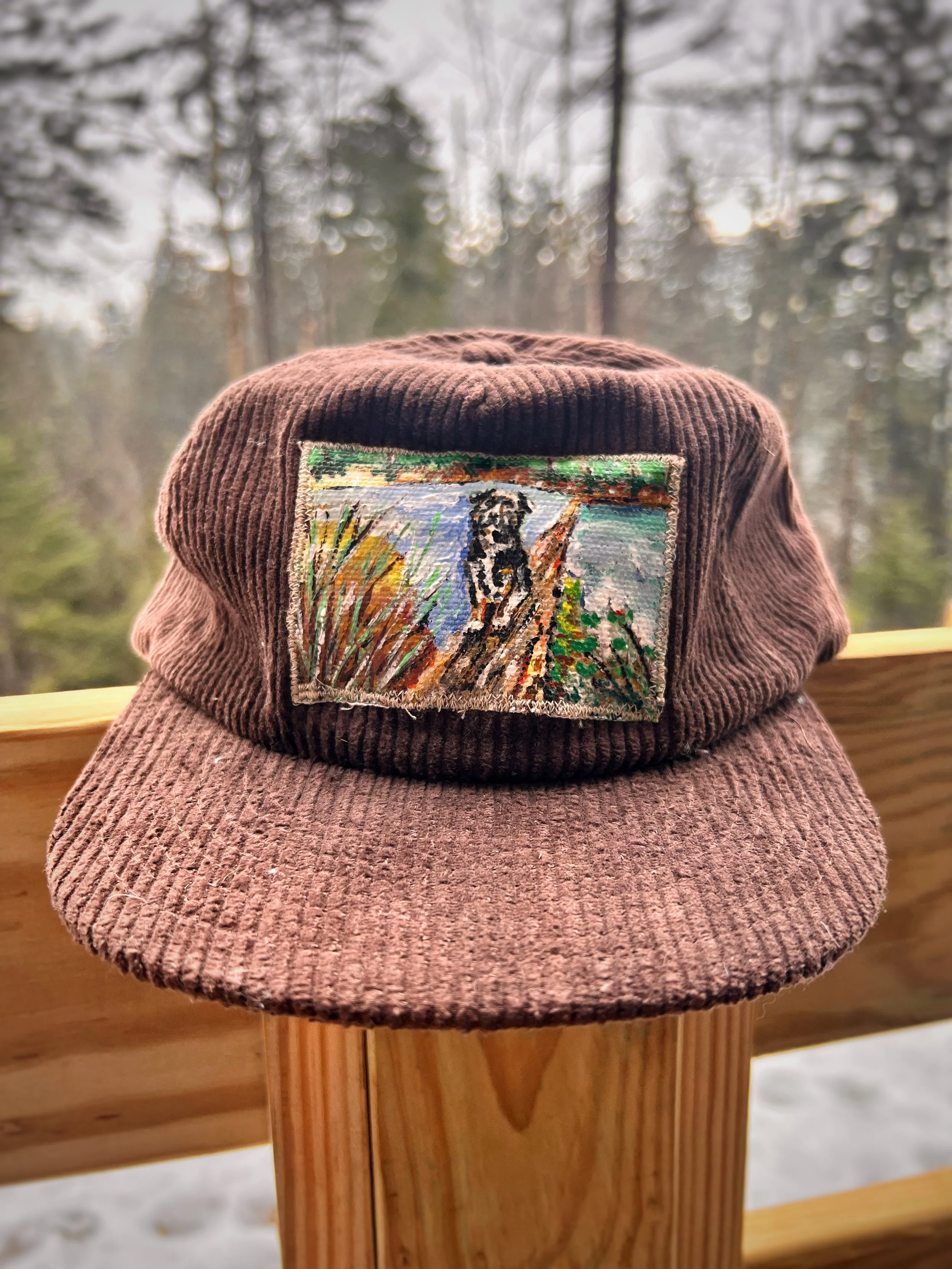 A brown corduroy cap with a rectangular embroidered patch of a black and white dog sitting on a log outdoors, with trees and sky in the background.