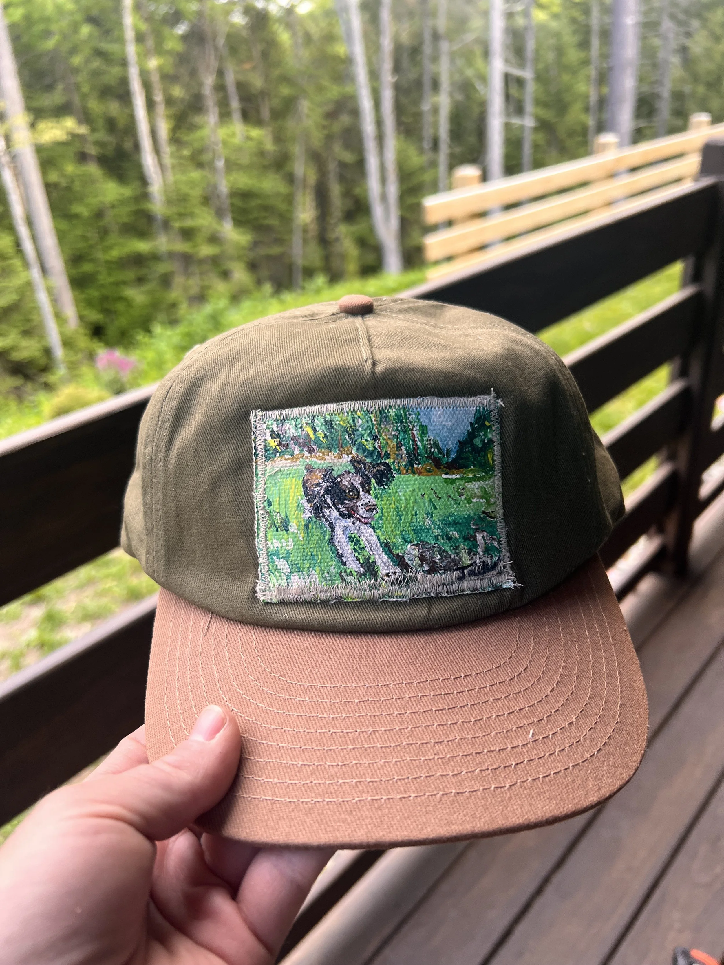 A cap with an embroidered patch of a dog and scenic background, held by a person's hand on a wooden deck with a forest in the background.