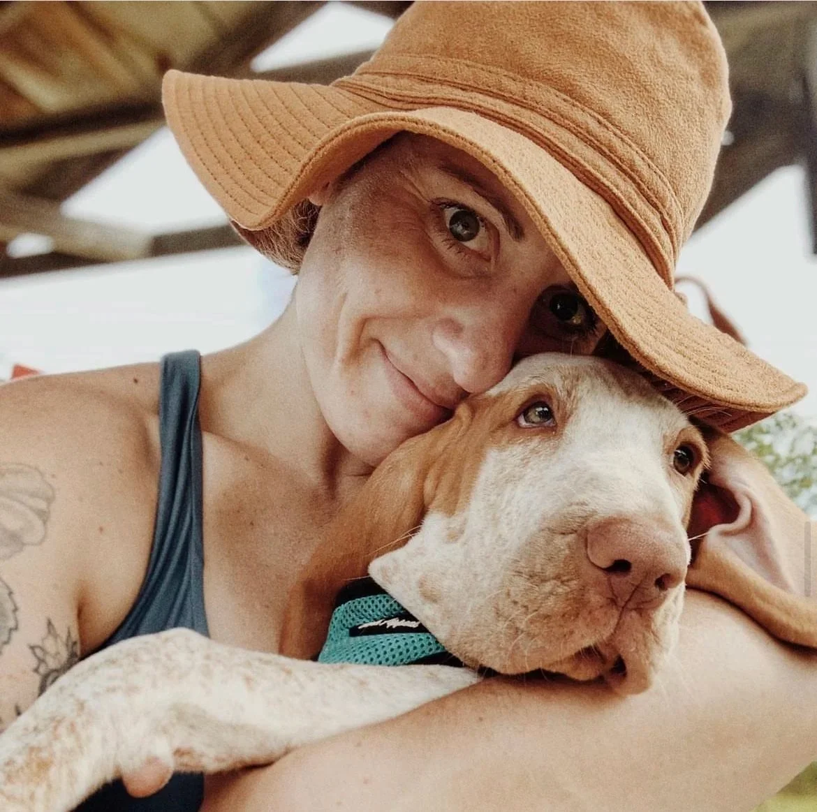 Woman wearing a wide-brimmed tan hat hugging a brown and white puppy.