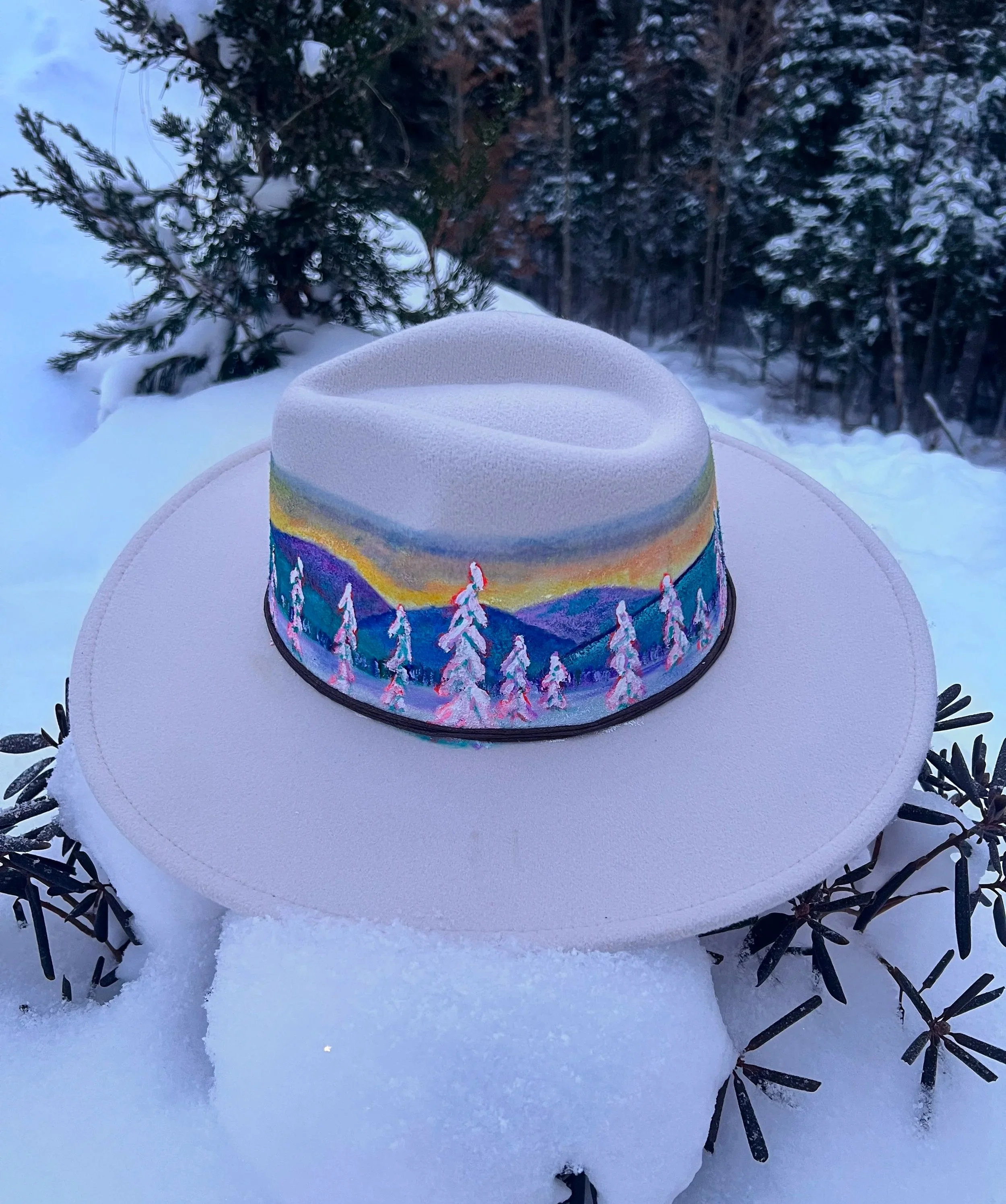 A white wide-brimmed hat with a colorful painted band depicting a mountain landscape and trees, resting on snow-covered branches outdoors in a winter setting with snow-covered trees in the background.