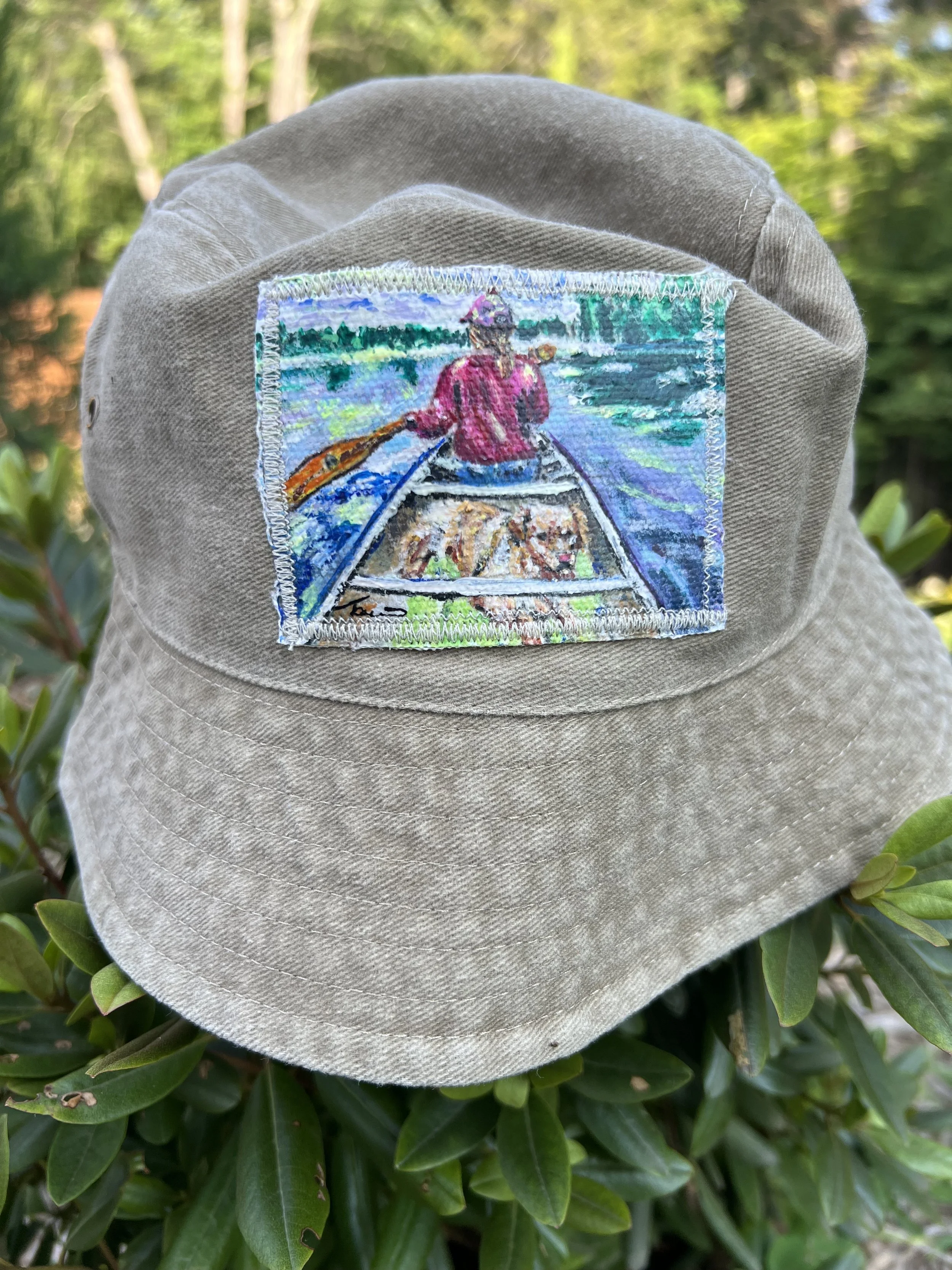 A beige bucket hat with an embroidered patch of a person rowing a boat on a lake or river, with greenery and water in the background.