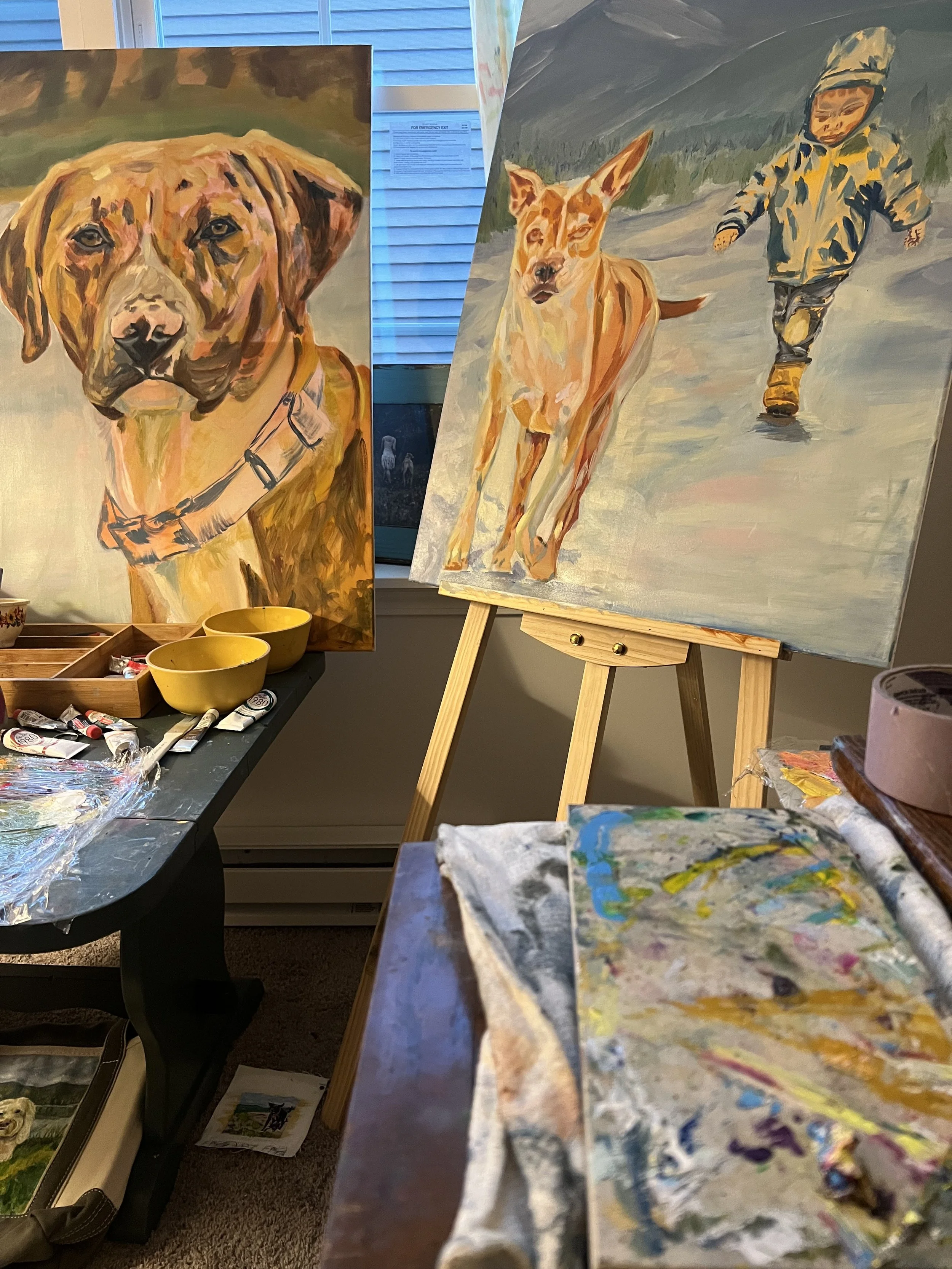 Two paintings of dogs in progress displayed on an easel and a wall, with painting supplies and a second artwork in the foreground.