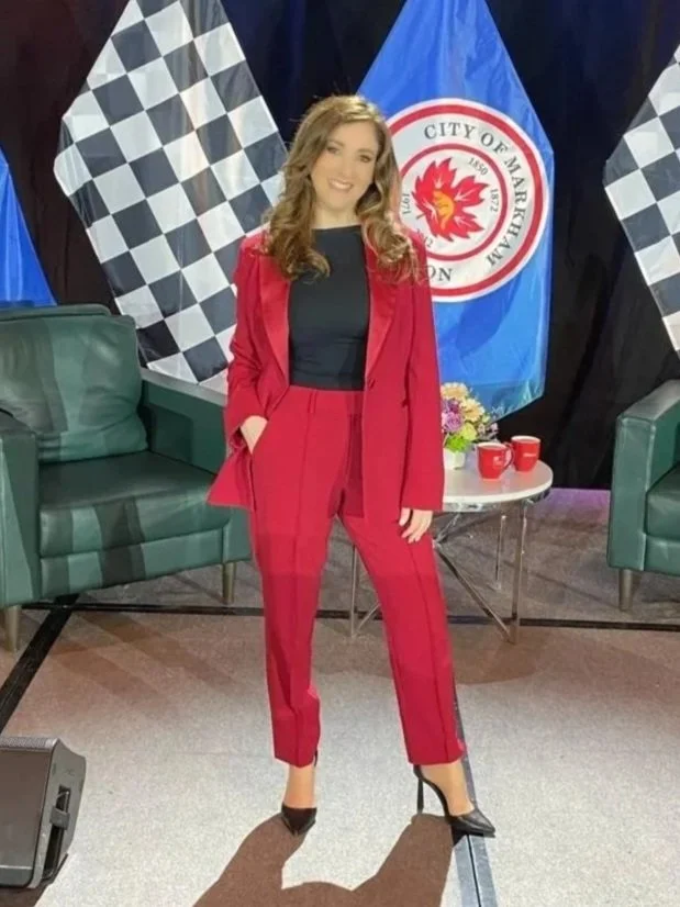Sonia Sache in a red suit and black top standing in a studio with green chairs, a small table with two red cups, a flower arrangement, and blue flags with a logo behind her.