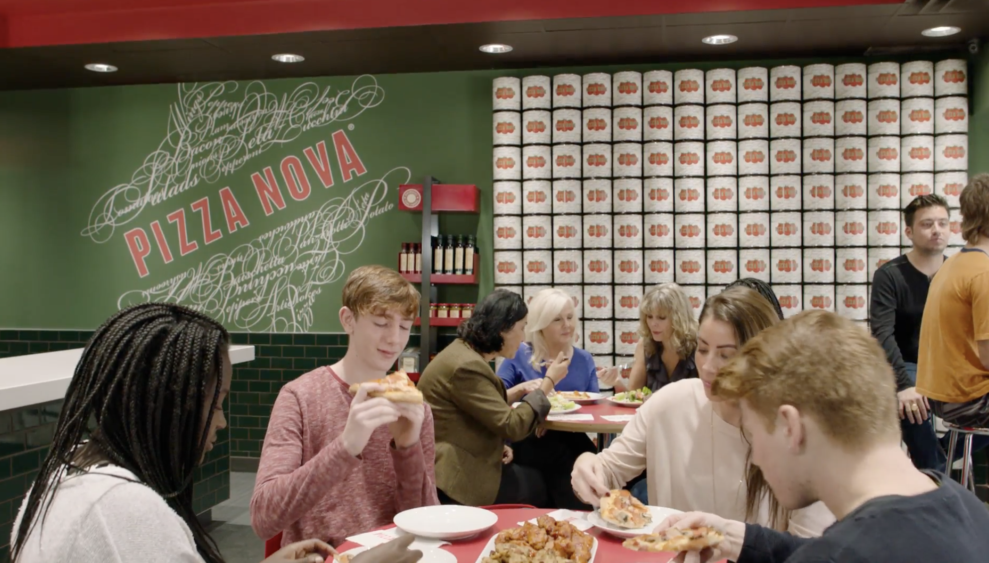 People eating pizza at a restaurant called Pizza Nova, with a green wall and a wall of pizza boxes in the background.