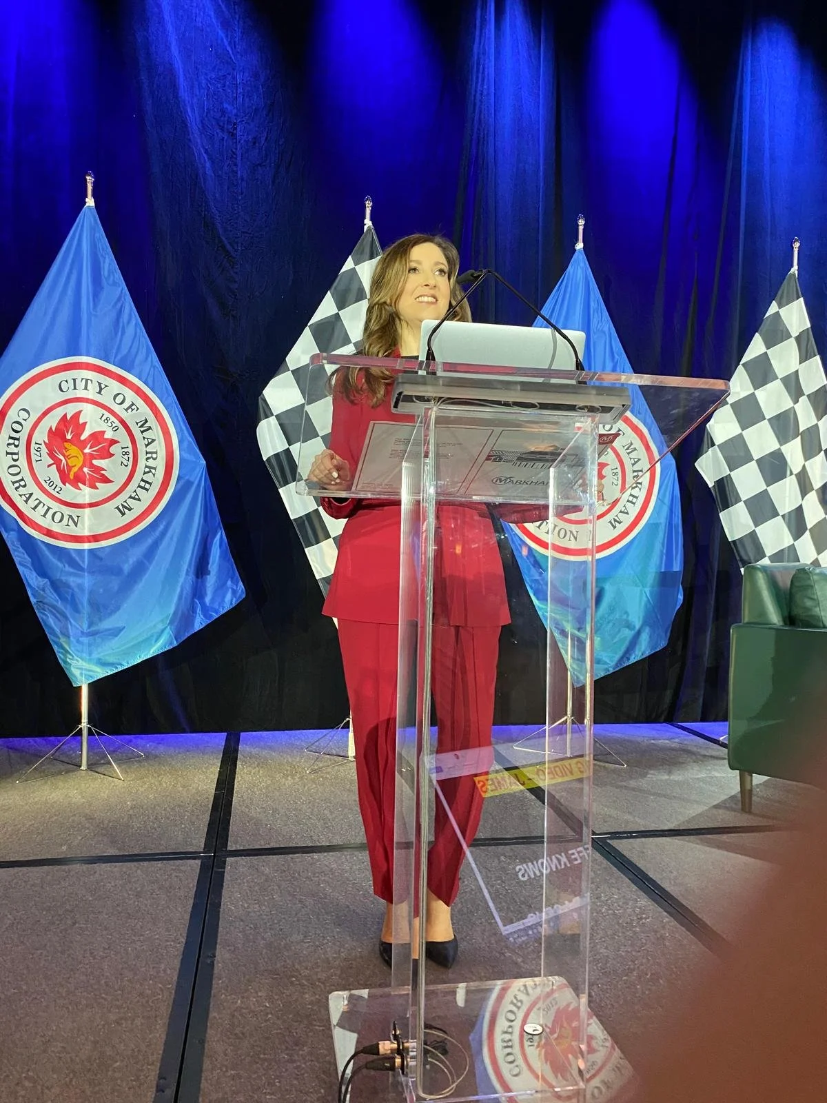 Sonia Sache in red suit speaking at a podium with several flags behind her, including city and checkered racing flags, on a stage with dark blue curtains.