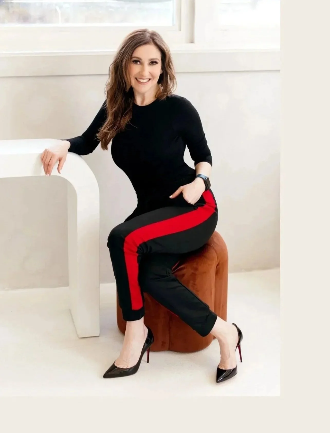 Sonia Sache is sitting on a brown cushioned stool, wearing black high heels, black pants with red and black stripes, and a black long-sleeve top, smiling at the camera in a bright room with white walls and a large window.
