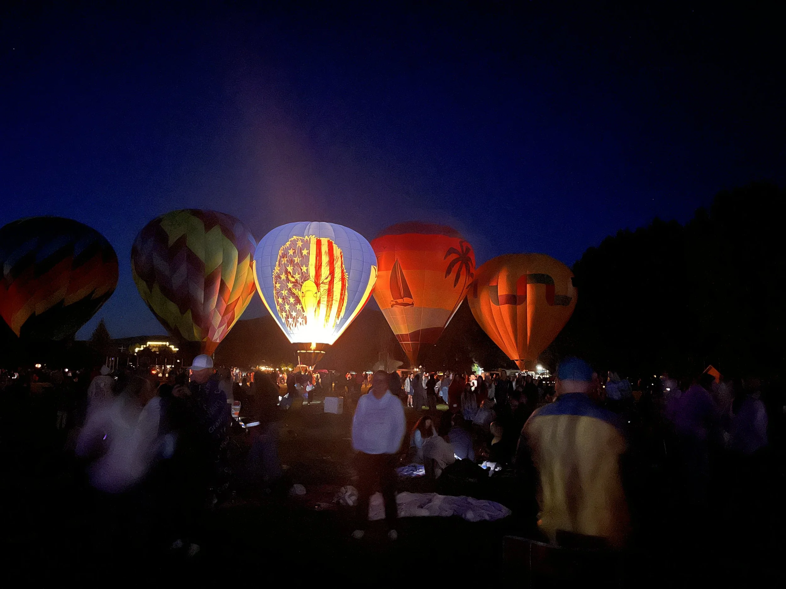 Nighttime scene of colorful hot air balloons, including one with an American flag design, floating above a crowd of people gathered on the ground.
