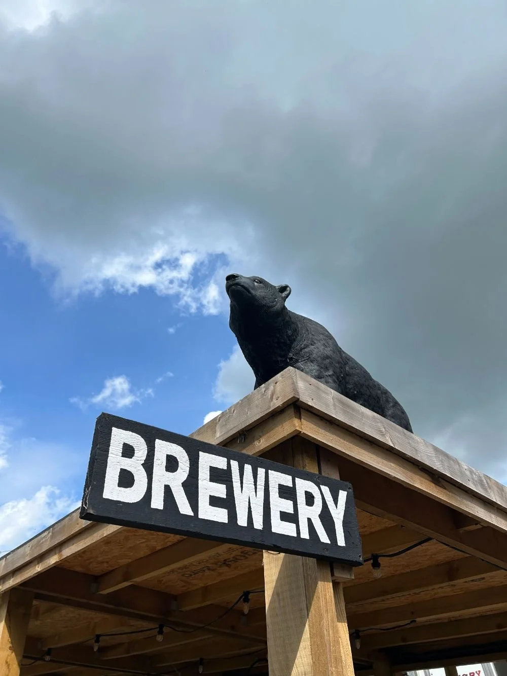 A black bear statue on a wooden platform with a sign that says 'BREWERY' in front of cloudy sky.