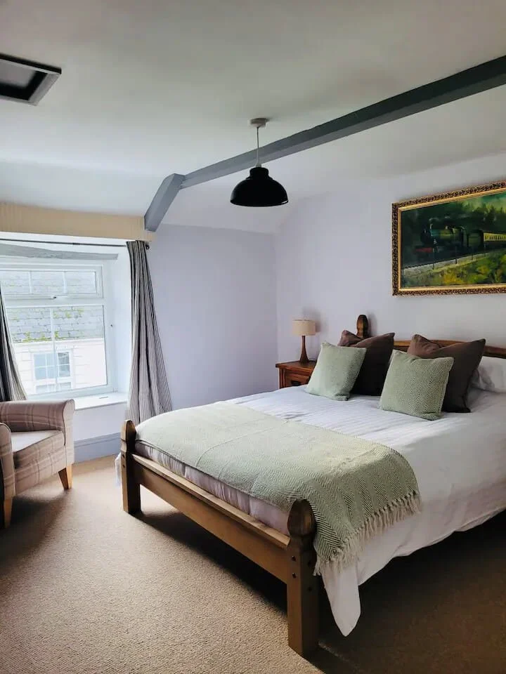 A cozy bedroom with a large bed, green and brown pillows, a green throw blanket, a wooden nightstand with a lamp, a framed landscape painting, a checkered armchair near a window with striped curtains, and a black ceiling pendant light.