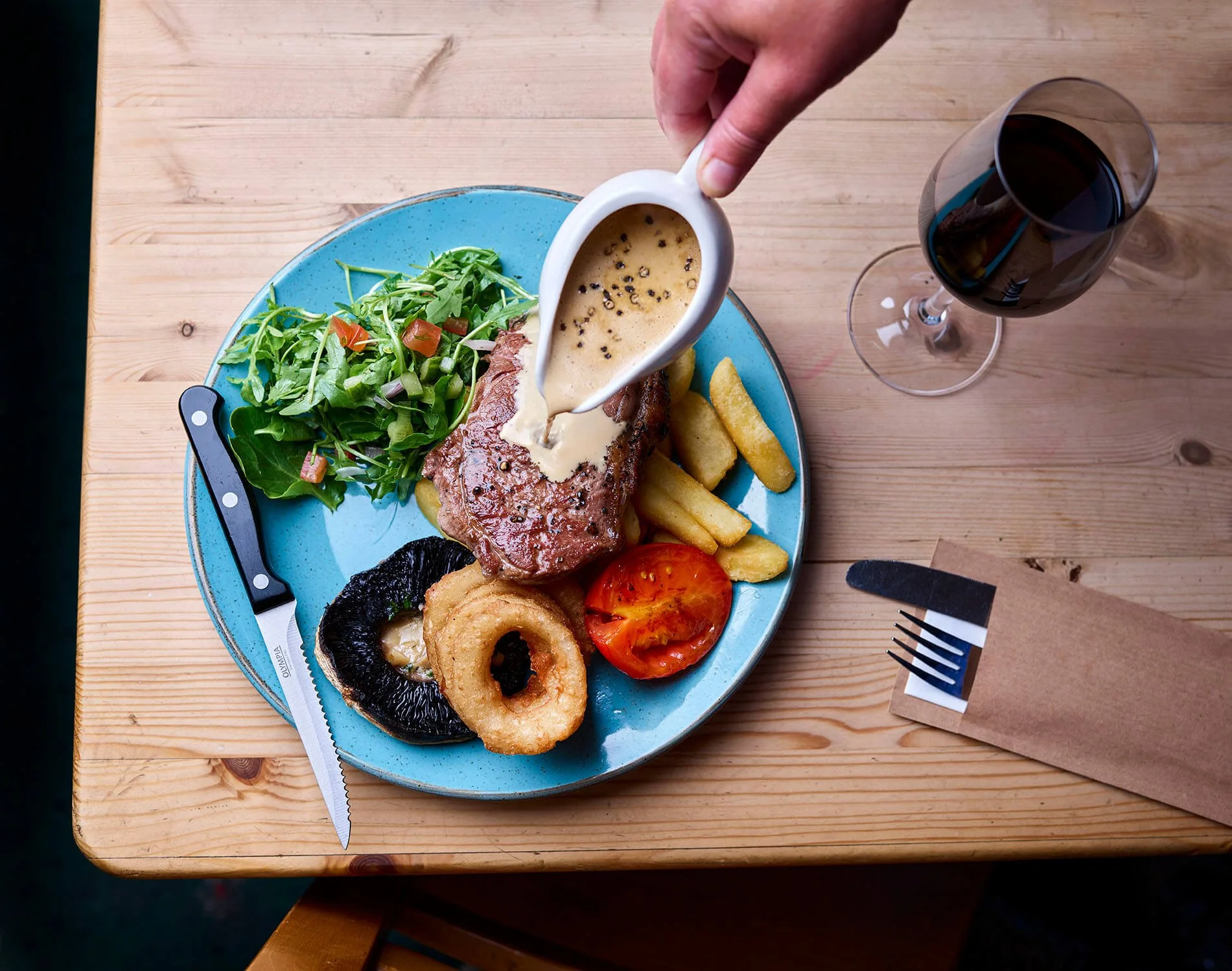 A dinner plate with a salad, steak topped with creamy sauce, onion rings, grilled tomato, and a black mushroom. A person's hand is pouring pepper sauce over the steak. There is a glass of red wine on the table. A knife and fork are placed on a paper napkin.