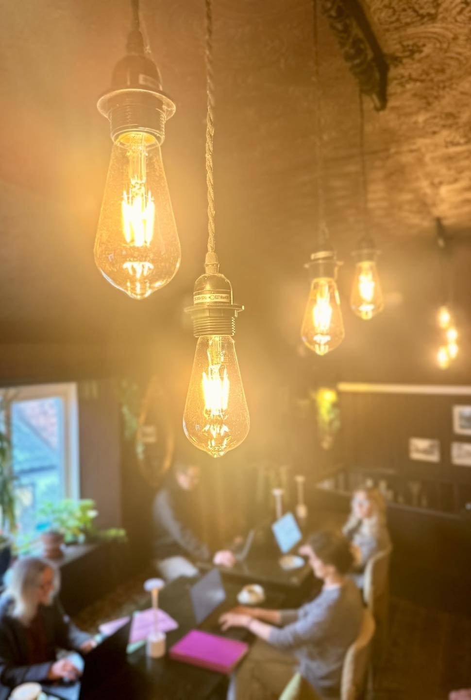 Close-up of hanging vintage Edison-style light bulbs emitting a warm glow in a dimly lit room with people working at a table in the background.