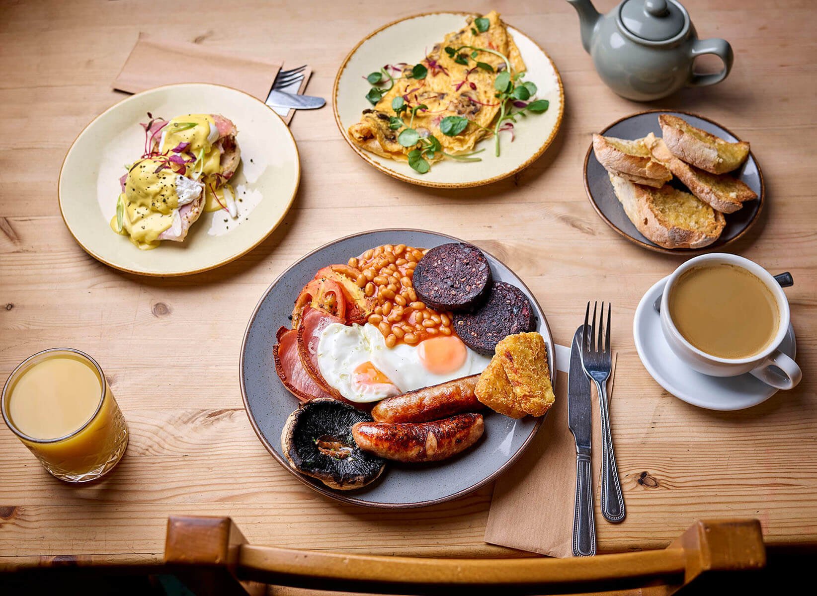 Breakfast meal on a wooden table, including eggs, sausages, bacon, baked beans, black pudding, grilled mushroom, fried bread, toast, orange juice, and tea.