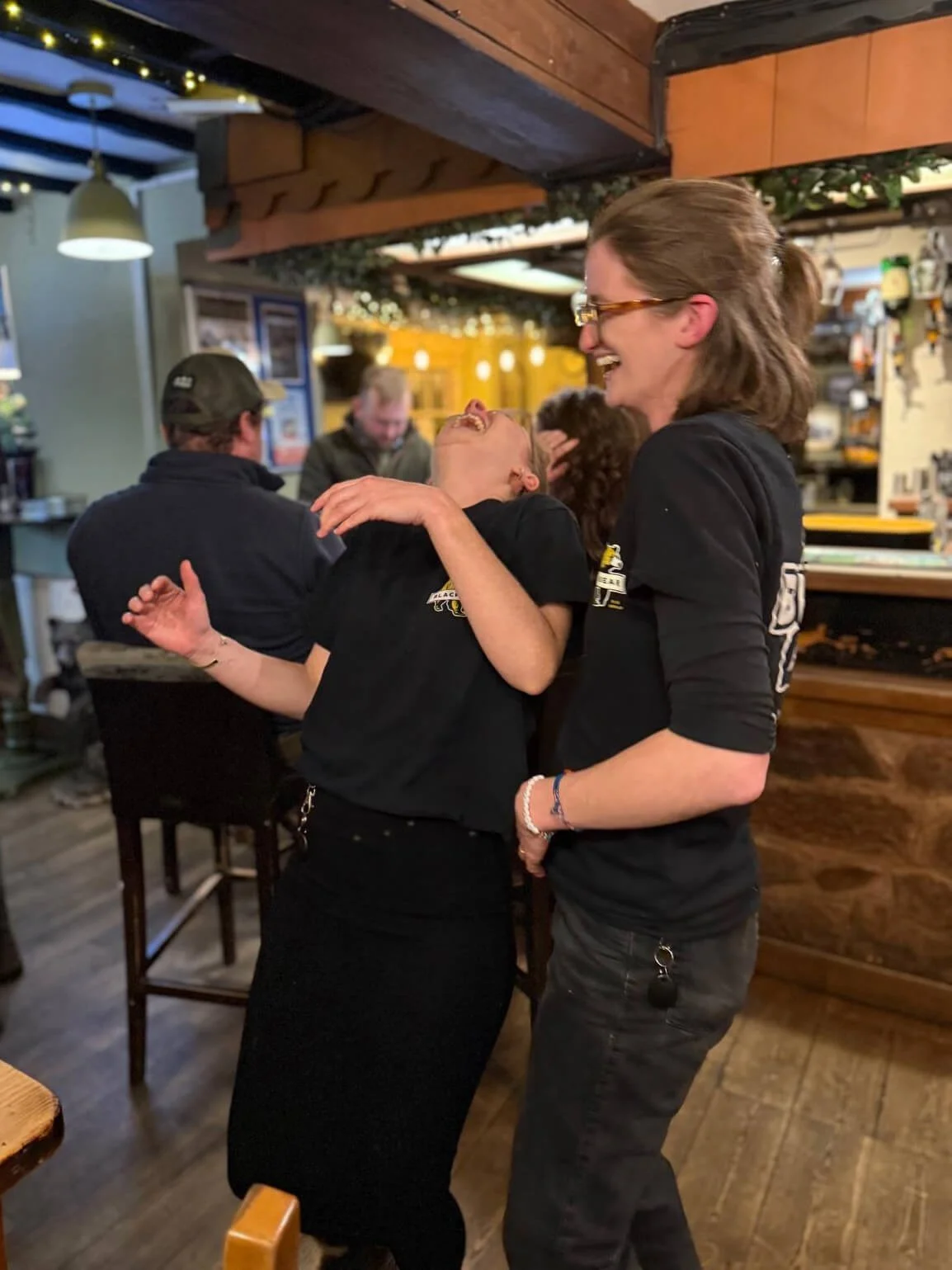 Two women are laughing and embracing in a bar or restaurant, with other people sitting and talking in the background.