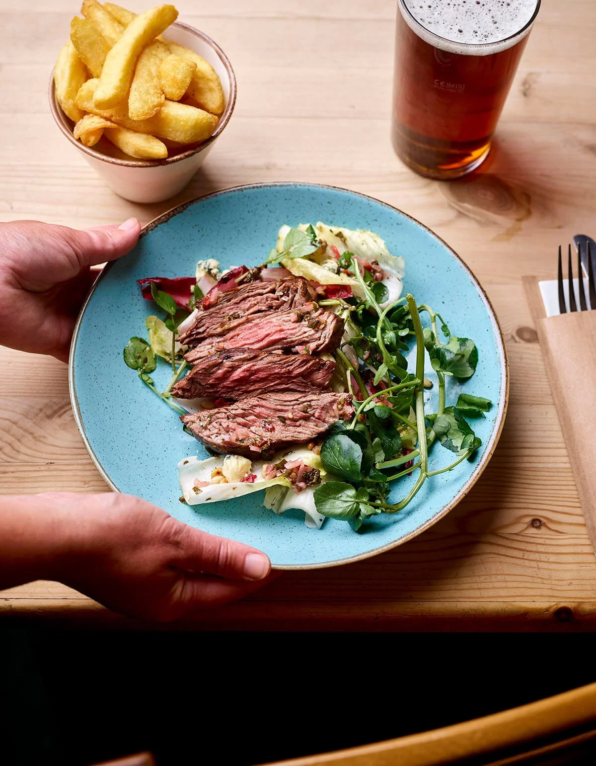A plate with sliced steak and salad greens, a small bowl of French fries, and a glass of beer on a wooden table.