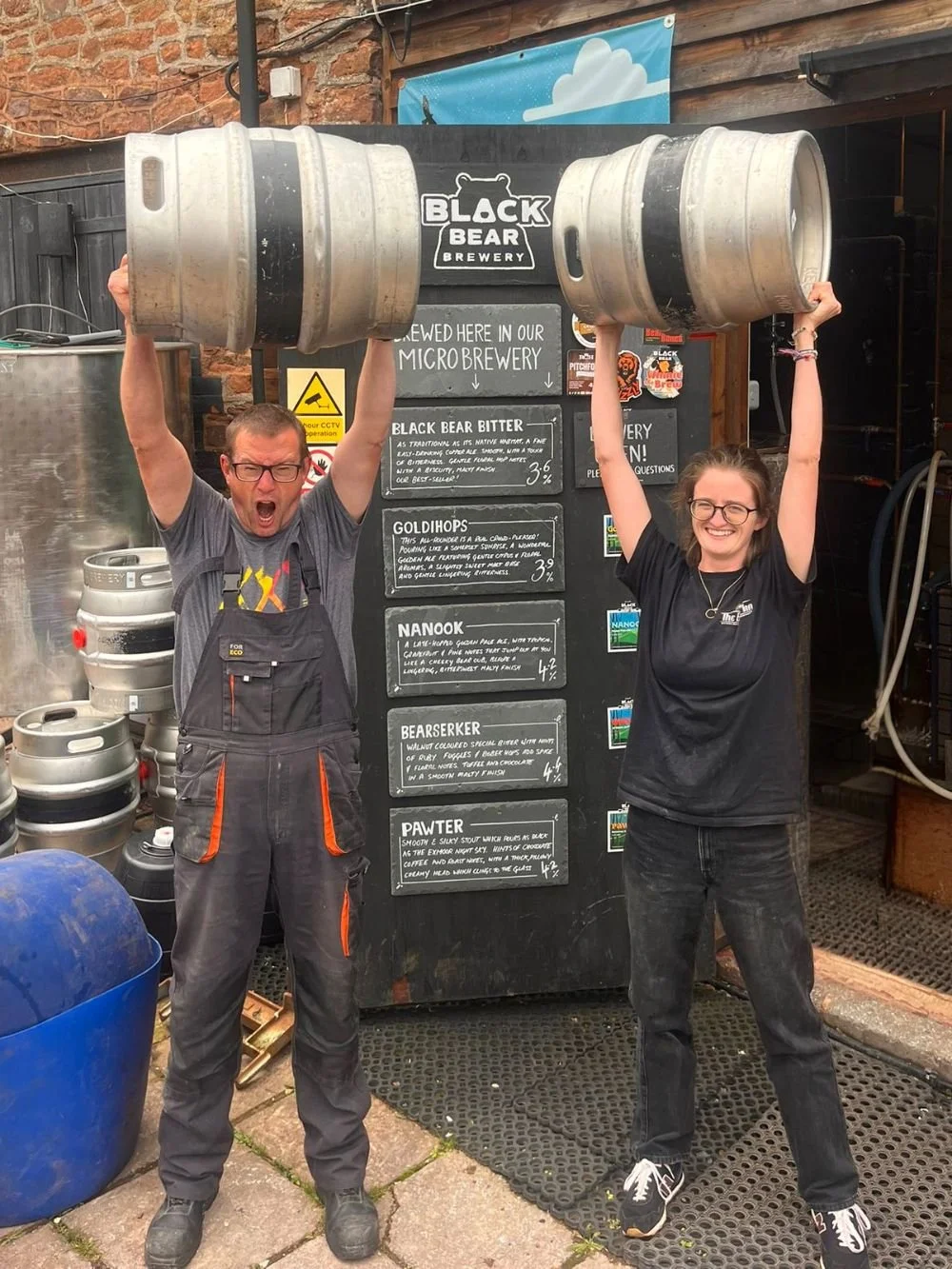 Two people standing each holding a large beer keg above their heads. The man on the left has glasses, is wearing a gray T-shirt and black overalls, and is shouting or cheering. The woman on the right has glasses, is wearing a black T-shirt and jeans, and is smiling. Behind them is a sign for Black Bear Brewery and a menu listing different beer options.