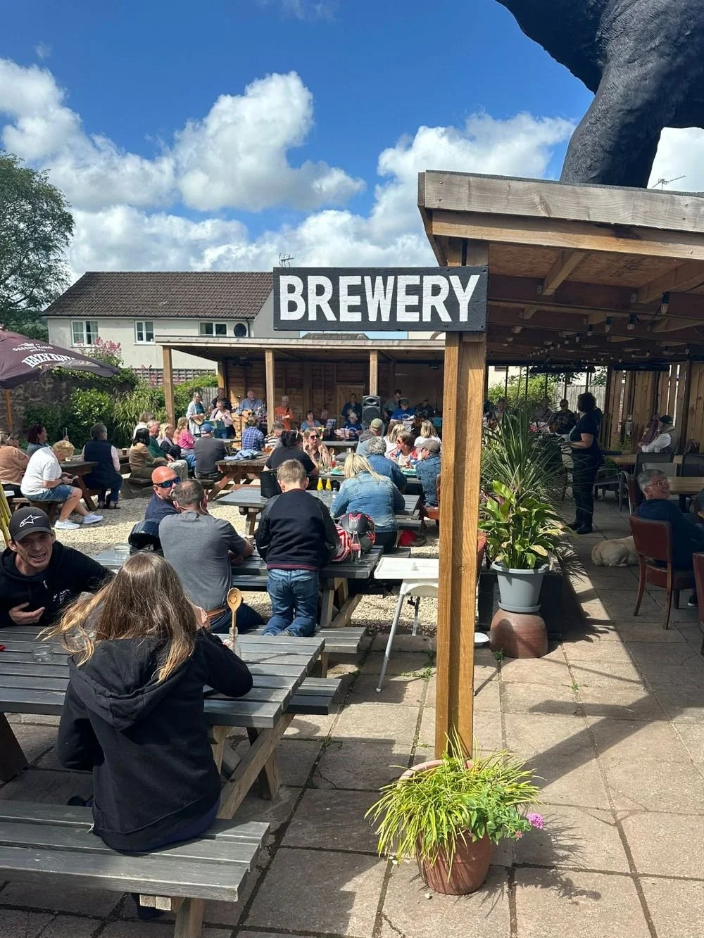 People gathered outdoors at The Bear Inn pub garden, sitting at wooden picnic tables under a wooden shelter, with a large sign reading 'BREWERY' in the foreground, potted plants, and a blue sky with clouds.