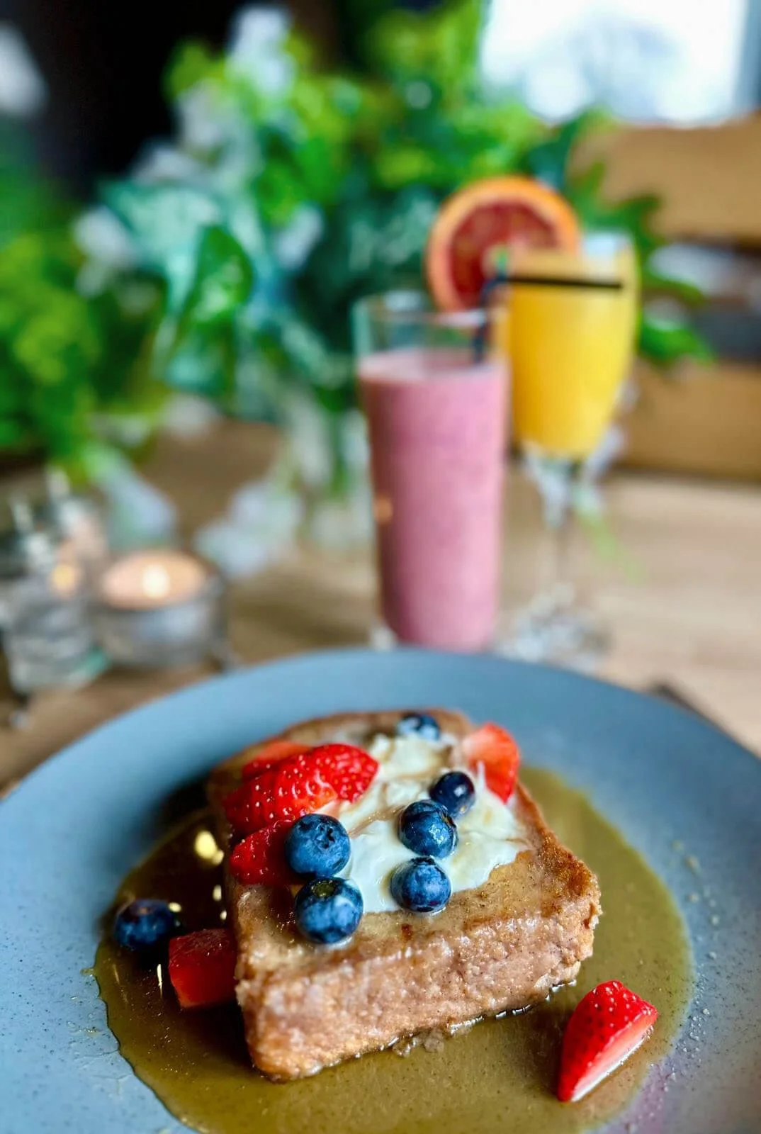 A dessert topped with strawberries and blueberries on a blue plate with a green sauce, in the background two colorful smoothies and a sliced grapefruit, with green plants and candles.