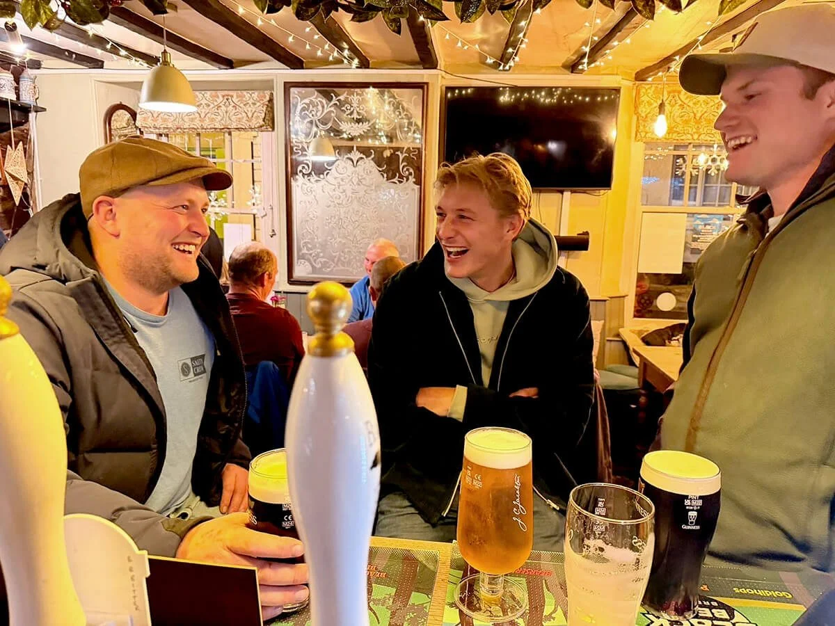 Three men are sitting at a bar table, laughing and enjoying drinks. The bar is decorated with string lights and has a cozy, warm atmosphere.