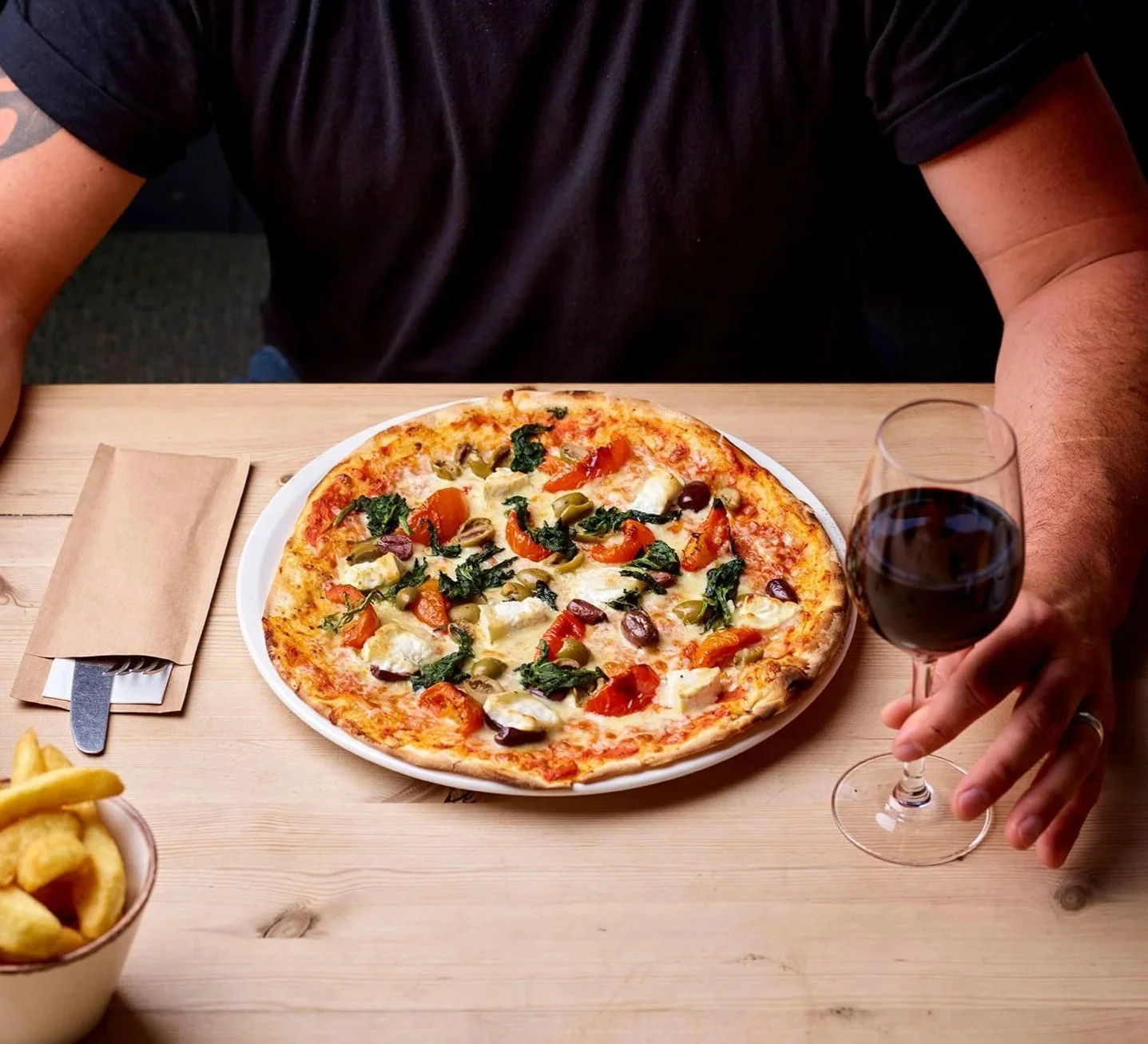 A person sitting at a wooden table with a vegetarian pizza topped with tomatoes, olives, and greens, and holding a glass of red wine.