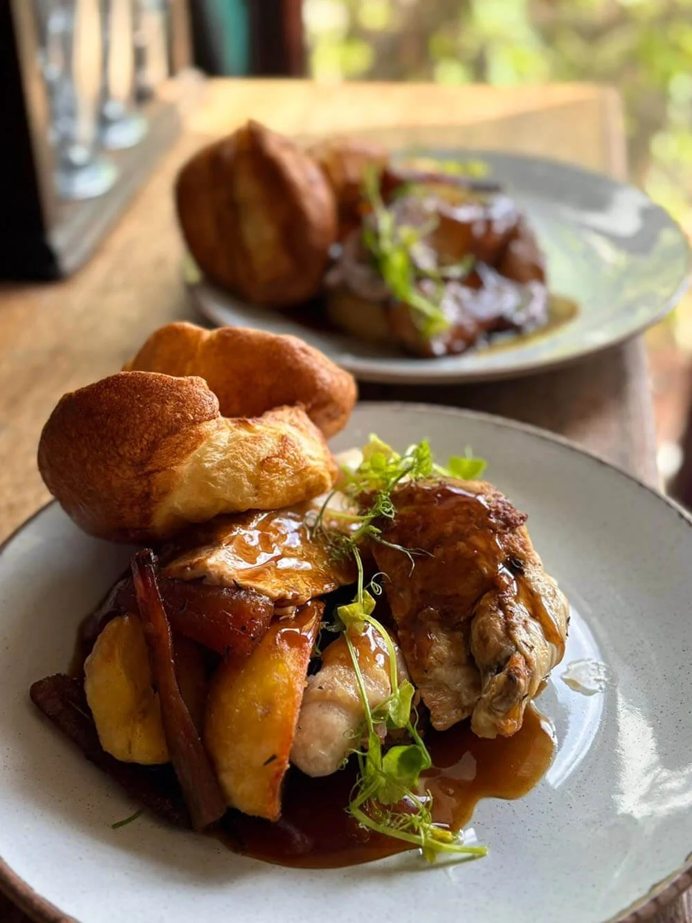 A plated serving of roast chicken with vegetables, two Yorkshire puddings, and garnished with green herbs in the foreground, with an additional plate of roast meat in the background.