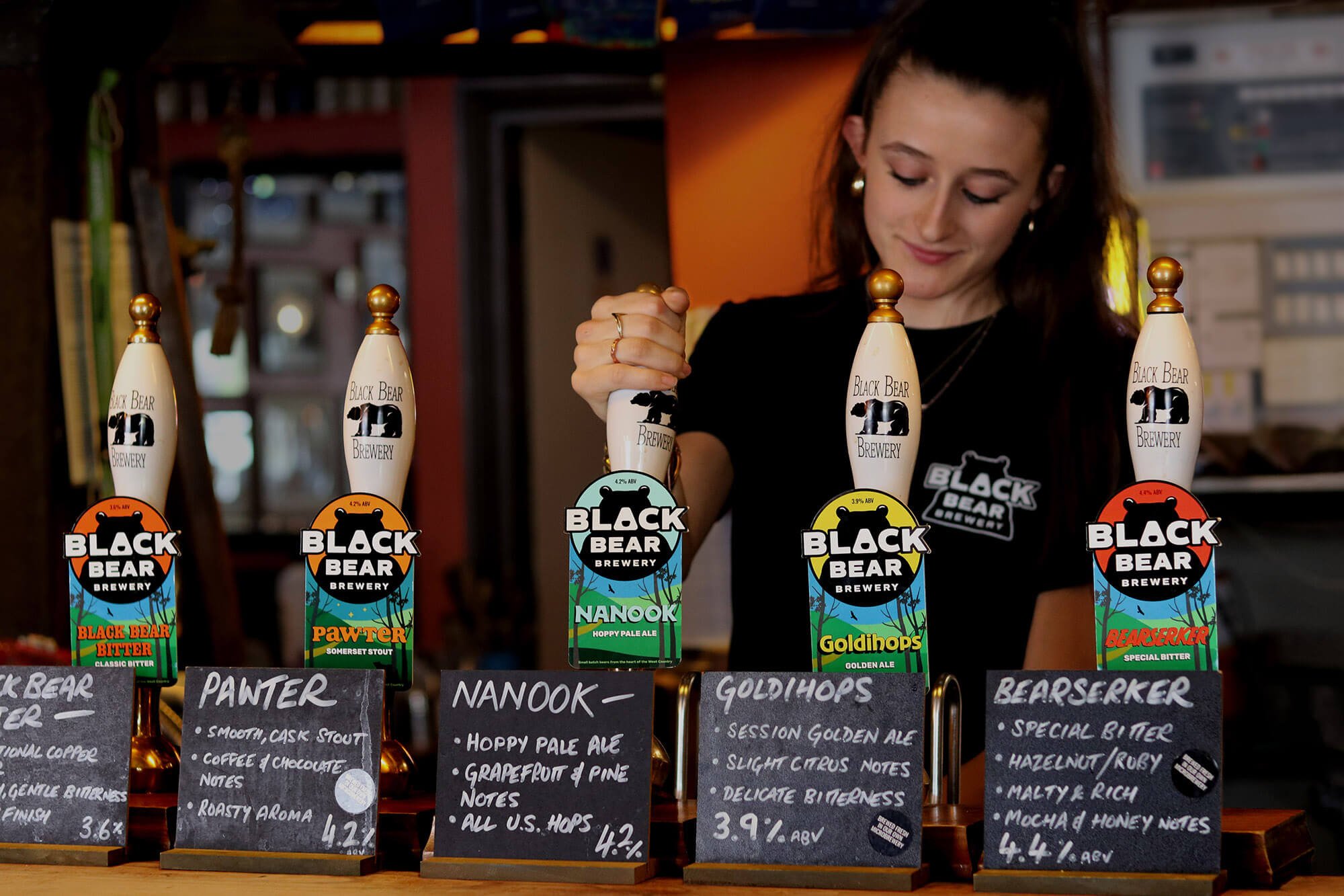 A woman with long dark hair standing behind a row of colourful Black Bear Brewery pump clips and handles, with handwritten chalkboard menus displaying beer descriptions and in the foreground.