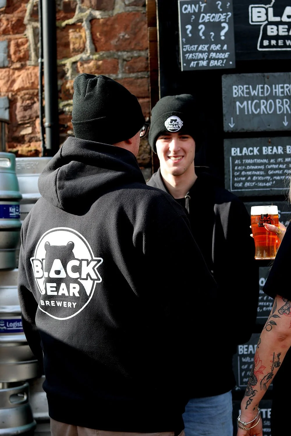 Two men with black beanies and hoodies with 'Black Bear Brewery' logos are talking at an outdoor bar. One man is holding a pint of beer.