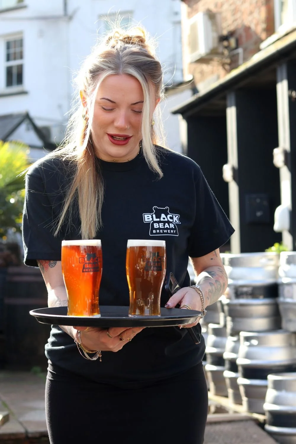 A woman with blonde hair and tattoos wearing a black Black Bear Brewery t-shirt, carrying a tray with two glasses of amber beer, outdoors near stacked metal kegs.