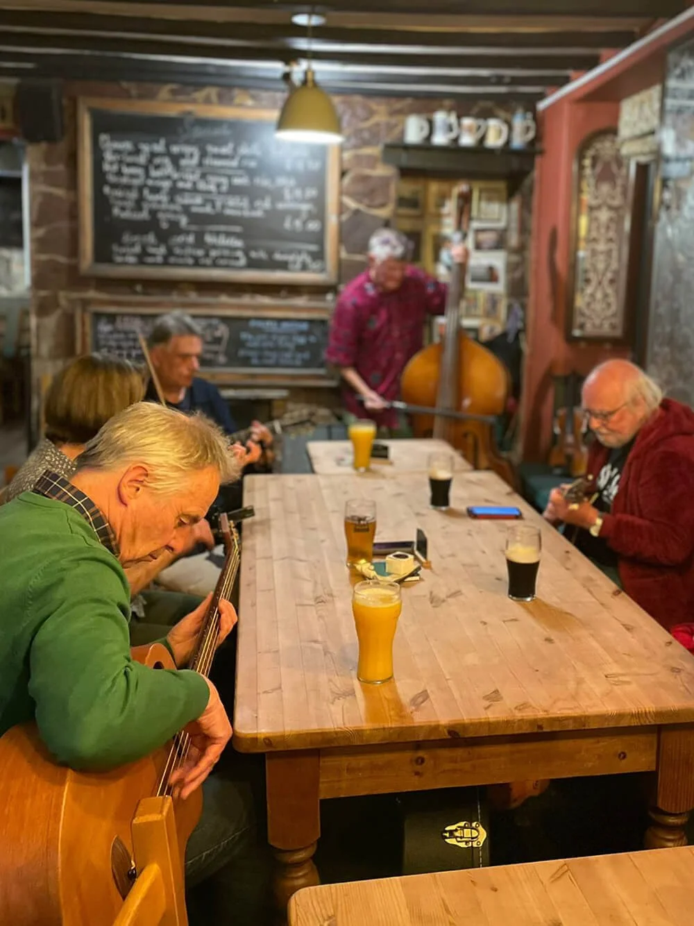 Group of people sitting around a wooden table in a rustic pub, some playing guitars, others drinking beers, with a musician in the background playing an upright bass.