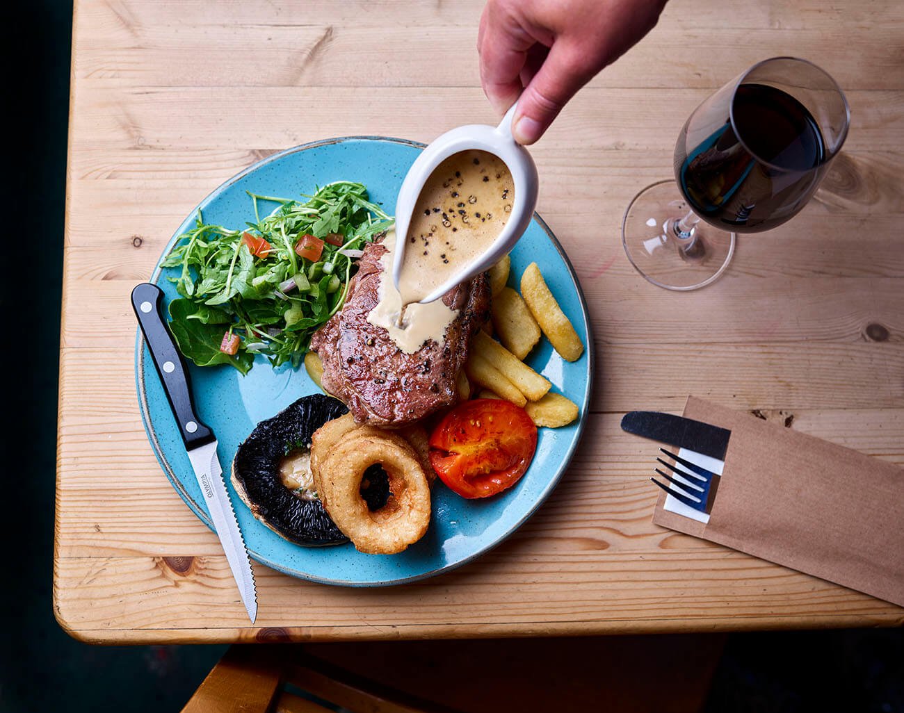 A plate of food with a salad, grilled steak topped with sauce, potato fries, a grilled tomato, and onion rings. A hand is pouring sauce over the steak, and there is a glass of red wine on a wooden table.