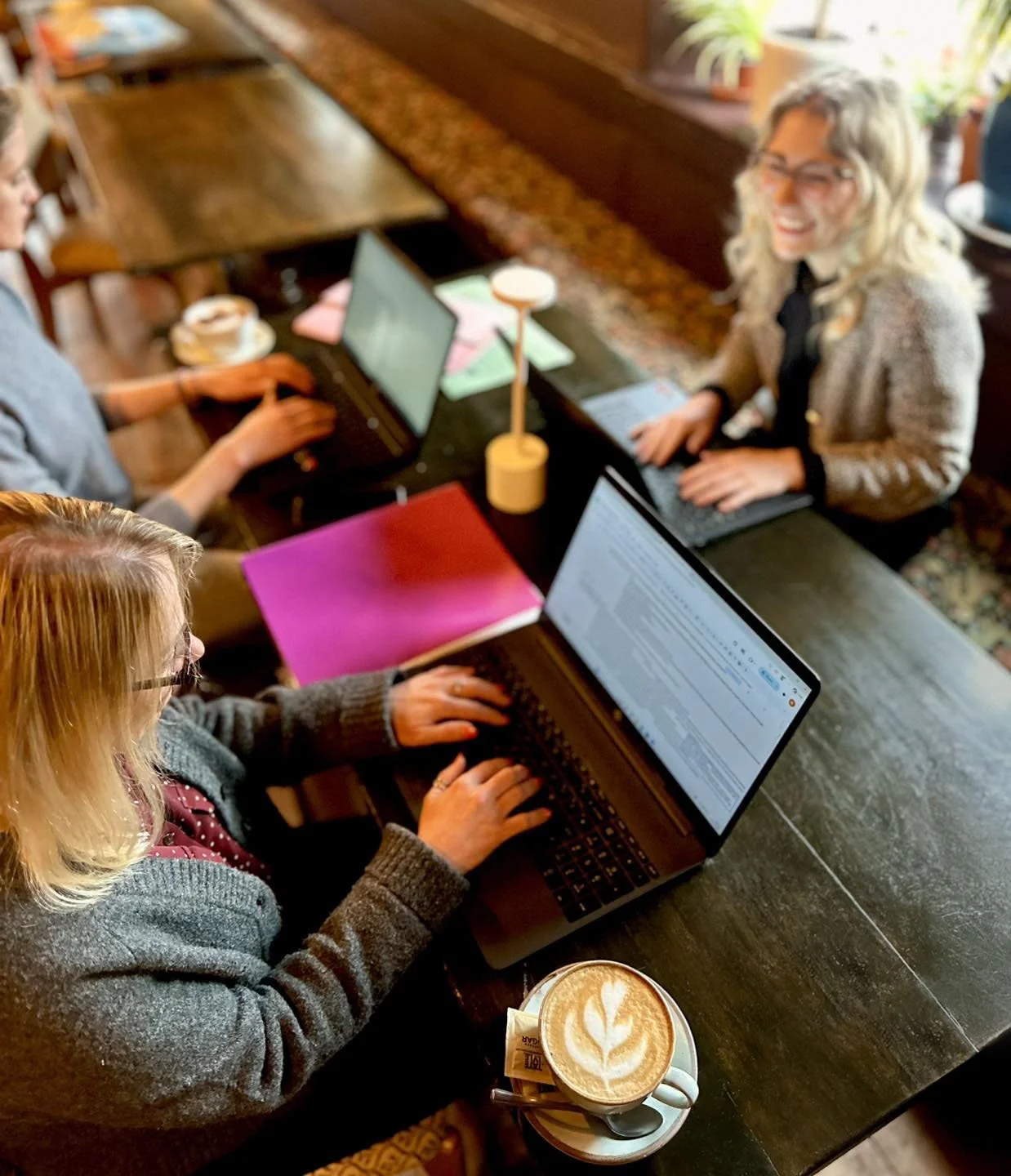 Four women working on laptops at a long wooden table in a cozy café with coffee drinks, notebooks, pens, and a decorative lamp. One woman has a cappuccino with latte art.