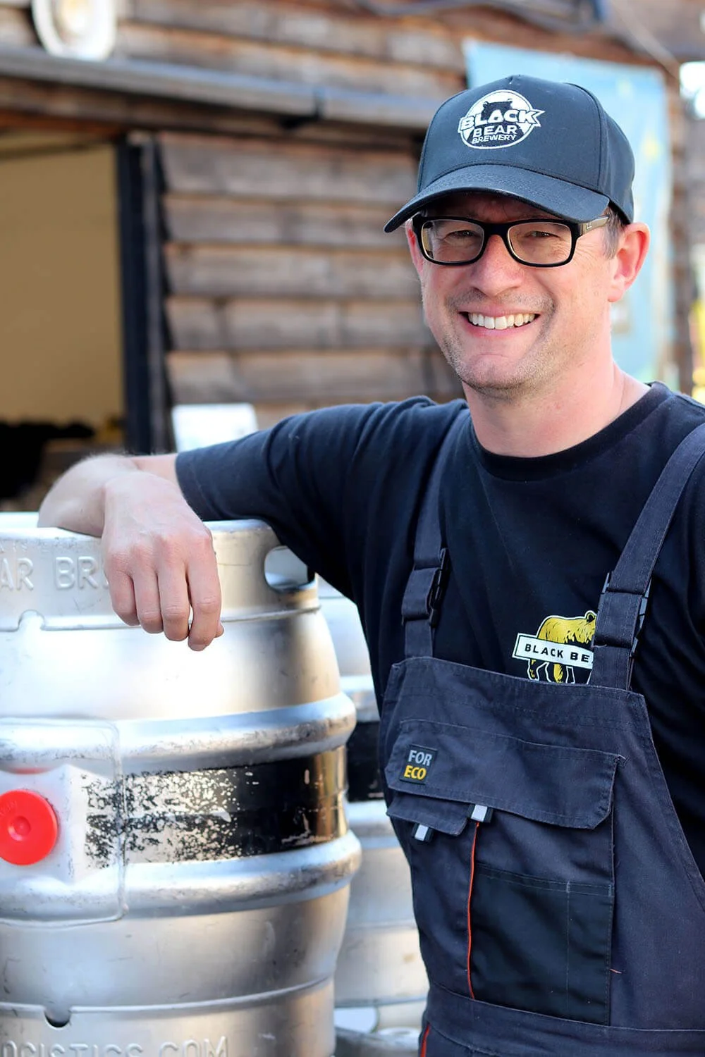 Head brewer Tom Davis, facing front, wearing glasses, smiling, wearing a black cap with the Black Bear Brewery logo, standing next to metal casks outside.