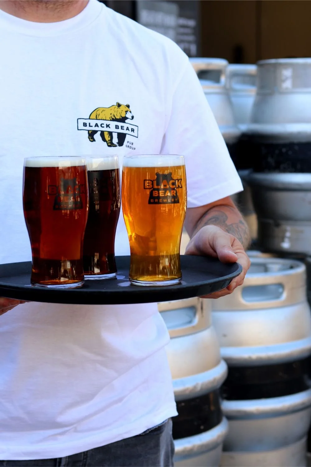 Person wearing a white t-shirt with a Black Bear logo, holding a tray with three glasses of beer in front of stacked beer casks.