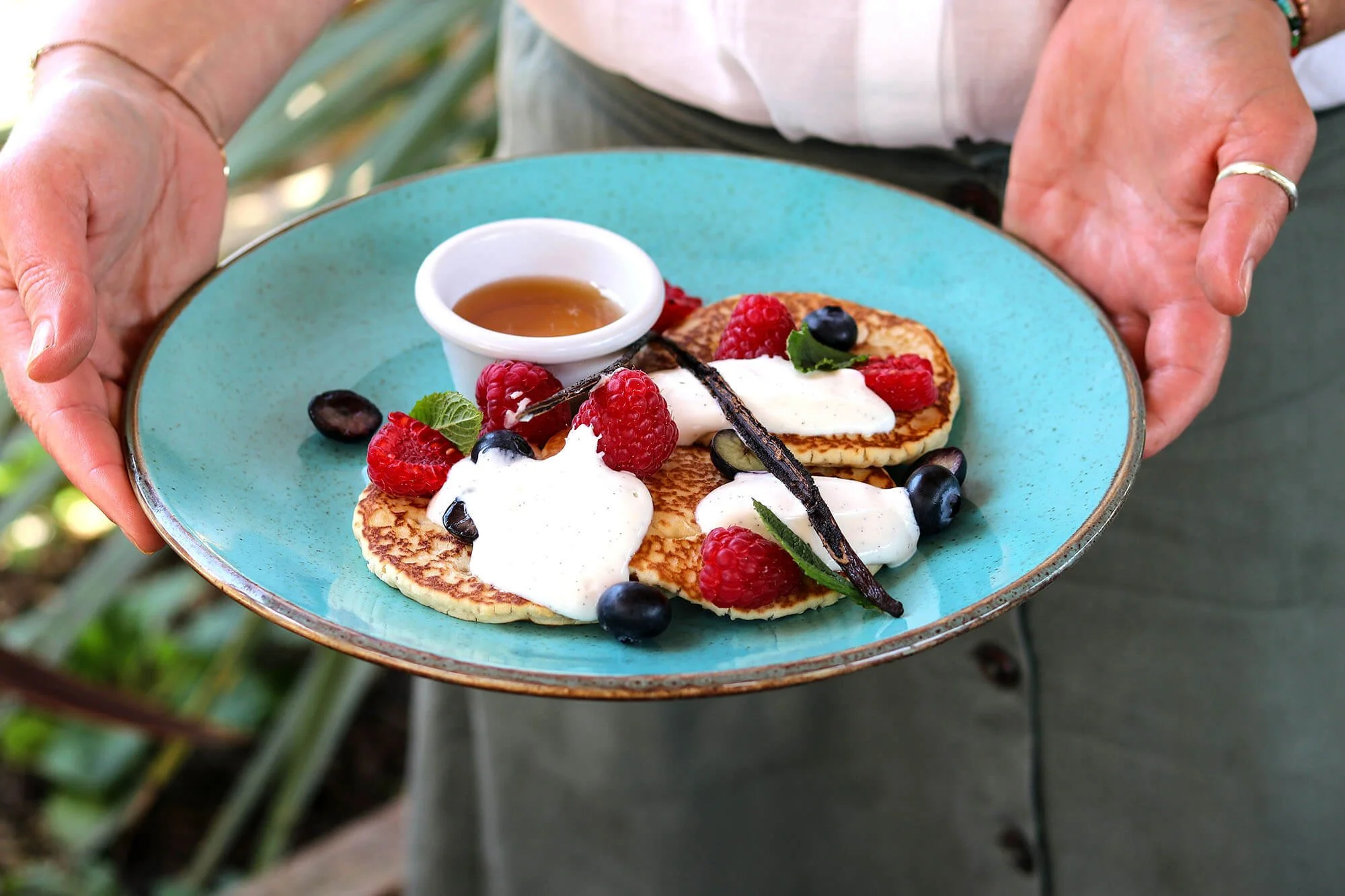 A person holding a turquoise plate with pancakes topped with raspberries, blueberries, whipped cream, mint leaves, a sauce, and chocolate drizzle.