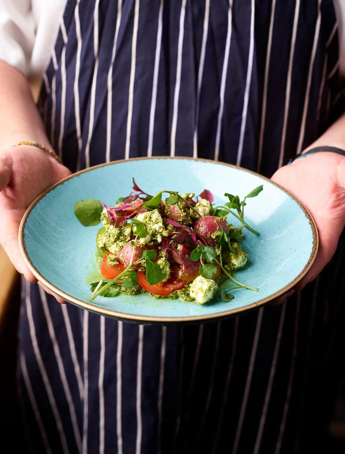 A person holding a blue plate with a salad containing cherry tomatoes, microgreens, and possibly cheese or crumbled features, garnished with herbs.