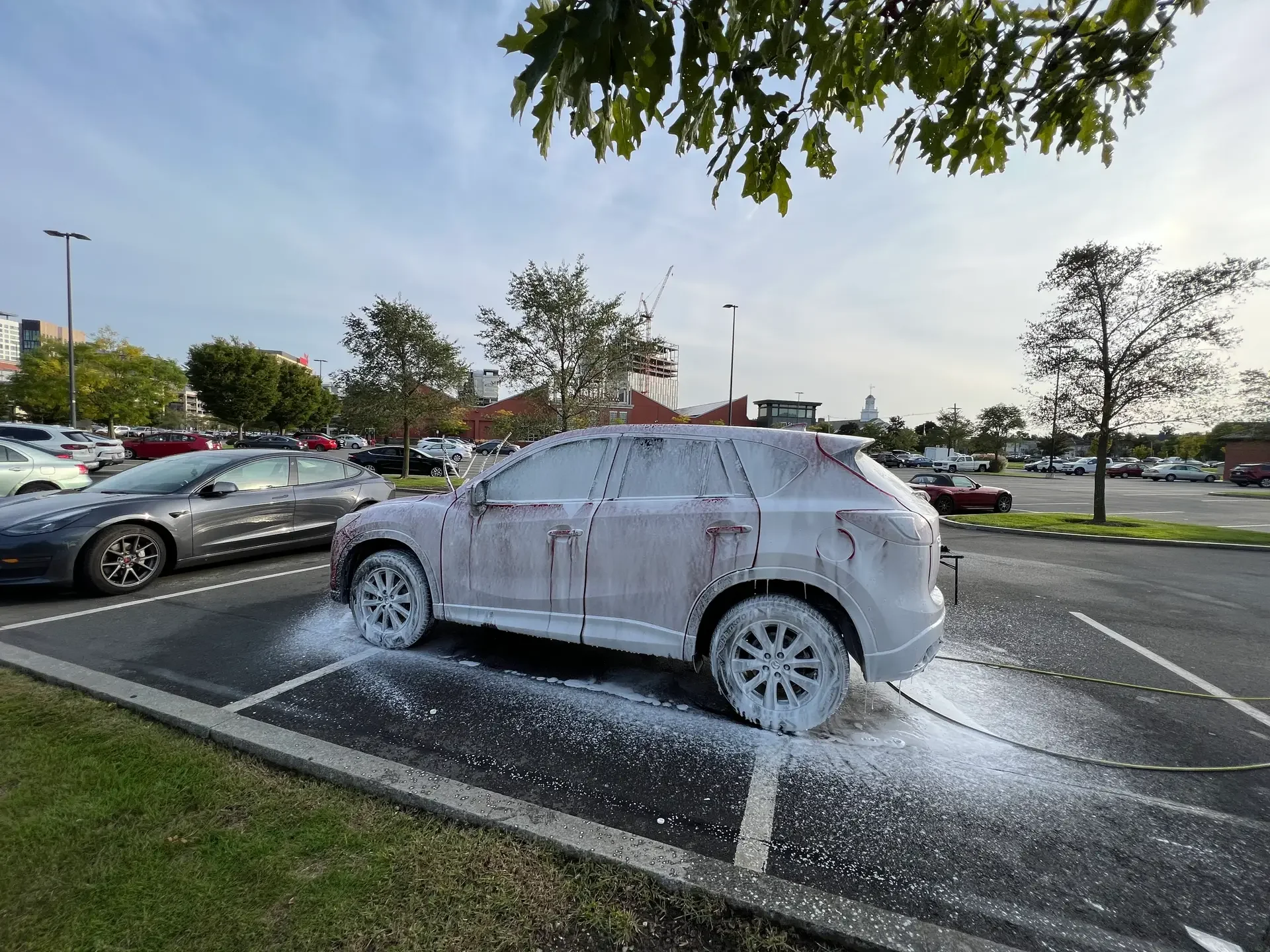 A car covered in soap foam during a wash in a parking lot, with other parked cars and trees in the background.