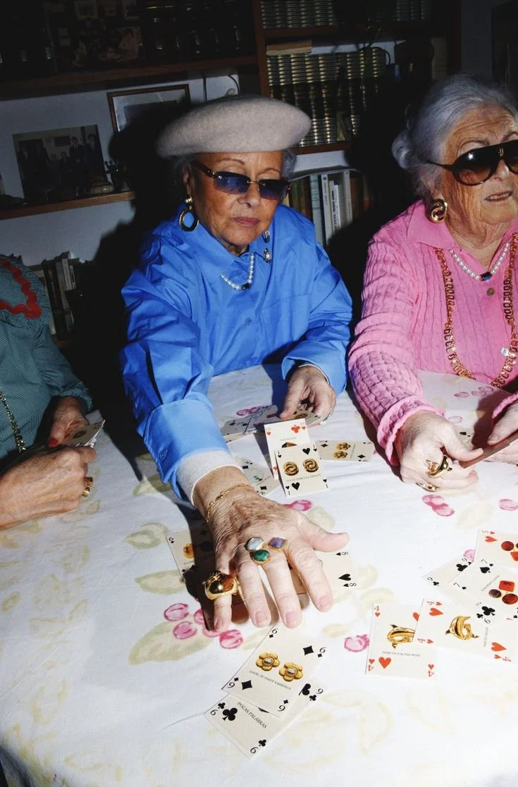 Groupe de femmes âgées jouant à un jeu de cartes, assises autour d'une table avec une nappe à motifs. Elles portent des vêtements colorés, des bijoux et des lunettes de soleil.