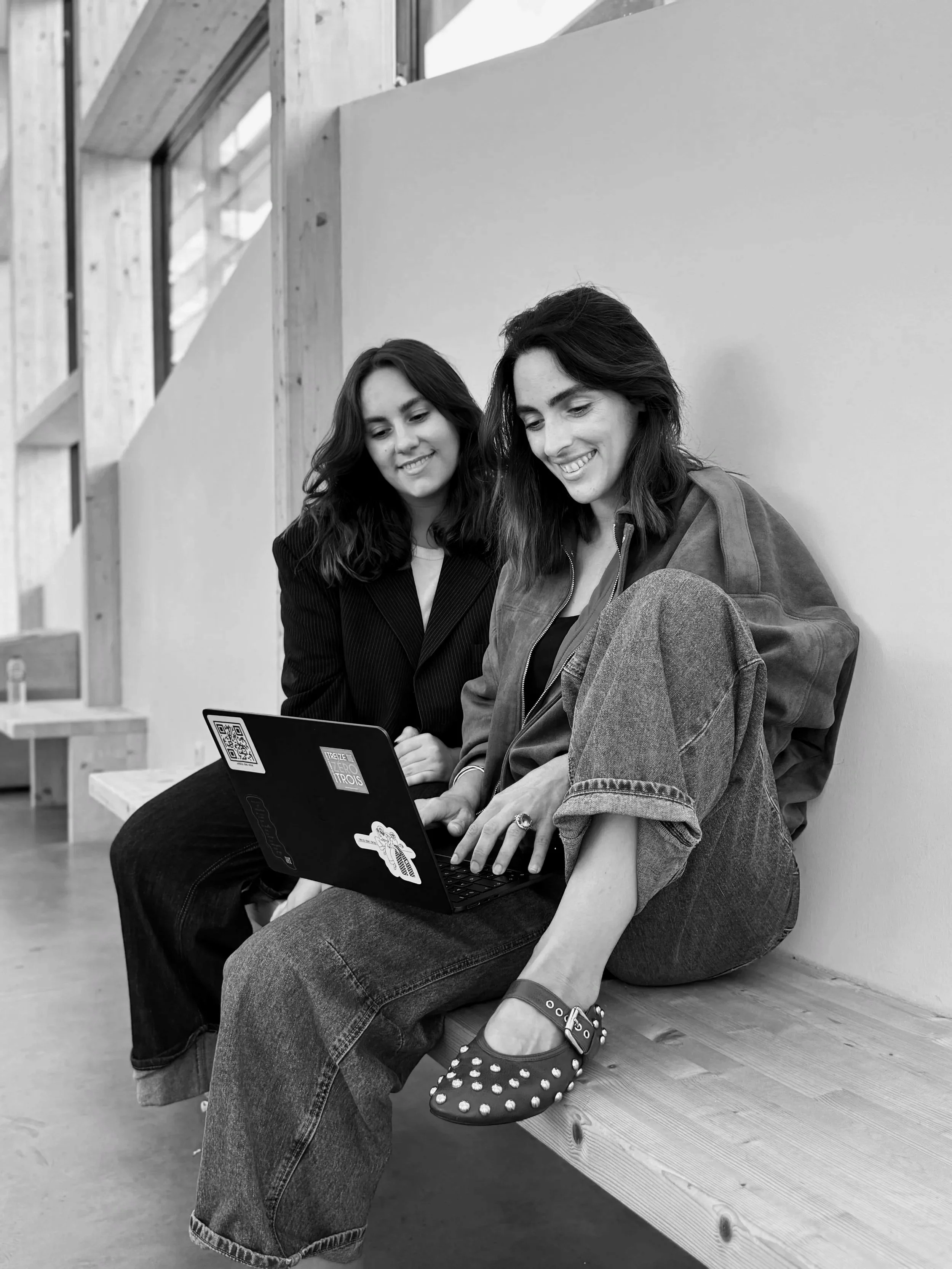 Emma et Alicia, les fondatrices de Treize Zéro Trois sont assises sur un banc en bois, regardant un ordinateur portable et souriant, dans un environnement moderne avec murs en bois et grandes fenêtres.