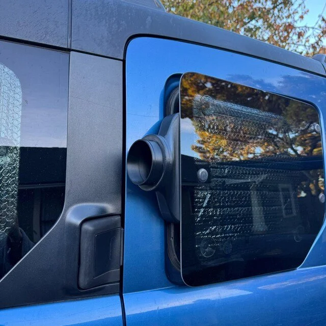 Close-up of the rear left side of a blue Jeep Wrangler, showing the rear corner, part of the door, and the rear window with reflections of trees and sky.