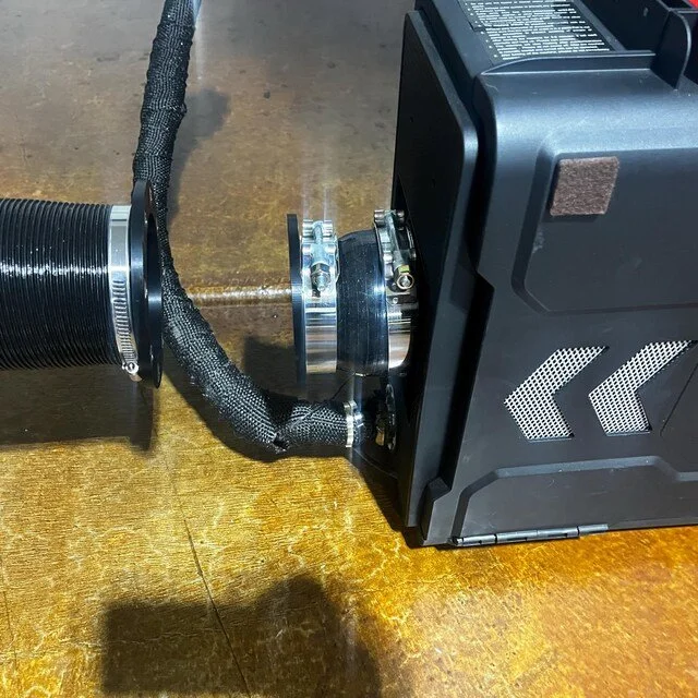 Close-up of a computer or electronic device connected to a flexible black hose with a metallic coupling, on a wooden surface.