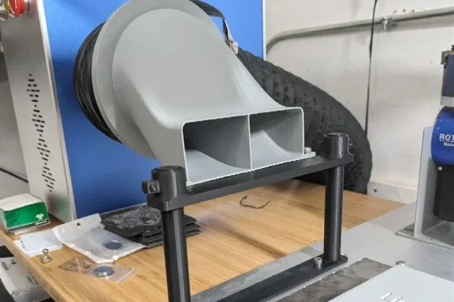 A laboratory device with a large gray and black funnel-like attachment mounted on a wooden table, with various small items and equipment nearby.