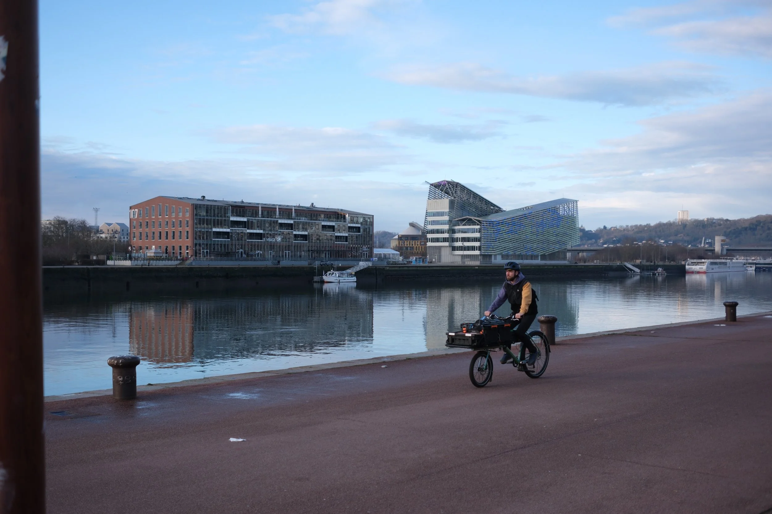 Un homme à vélo portant un casque, pédalant le long d'une rive de fleuve avec des bâtiments modernes en arrière-plan, sous un ciel partiellement nuageux.