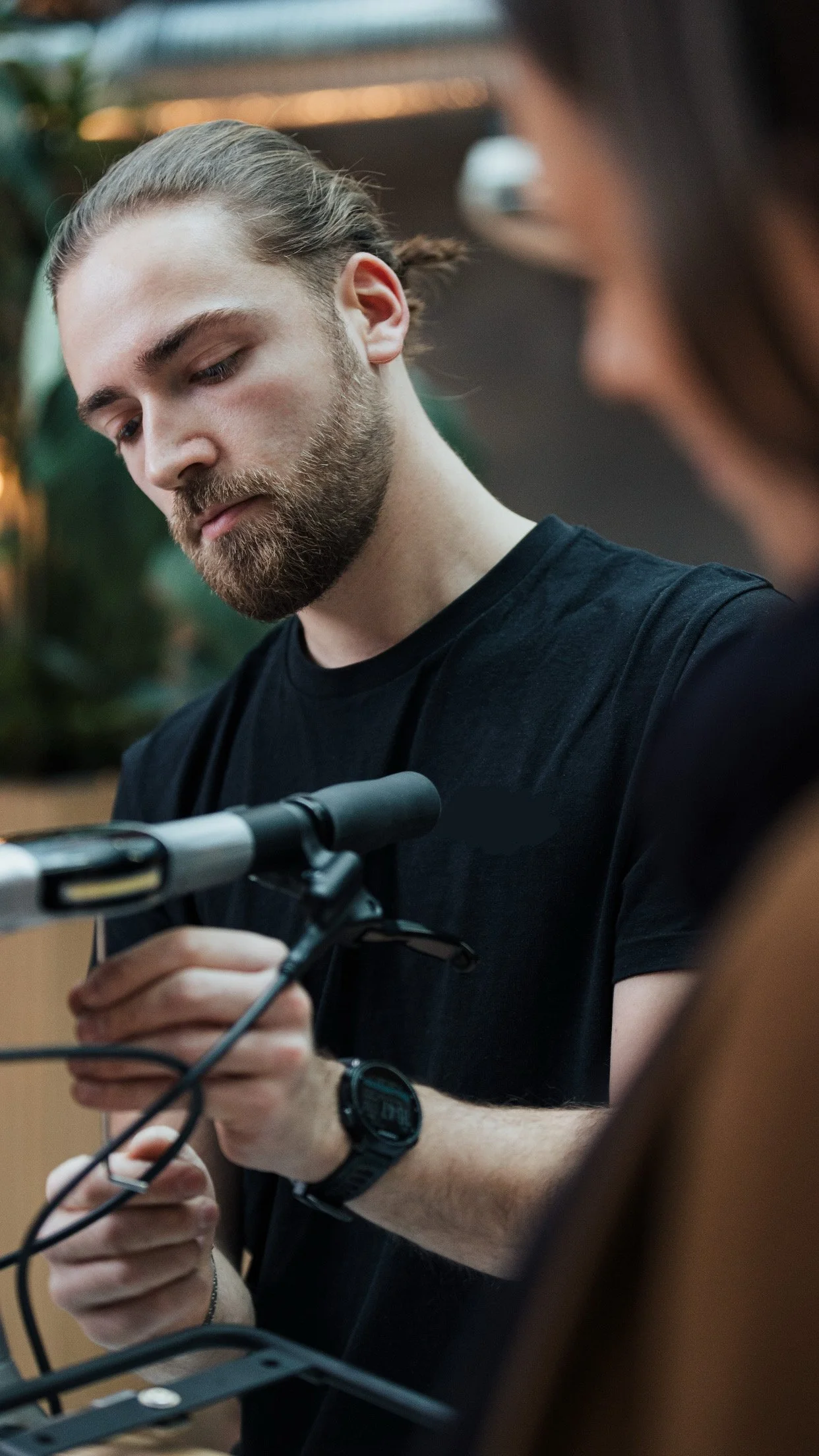 Un homme à barbe avec des cheveux longs attachés, portant un t-shirt noir, ajuste une micro sur un support, en intérieur avec un fond flou vert et chaud.